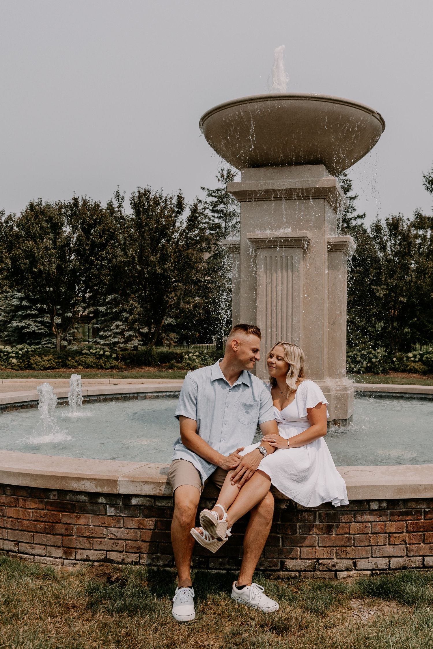A couple sits together on a brick ledge near a classic stone fountain in a romantic outdoor setting.