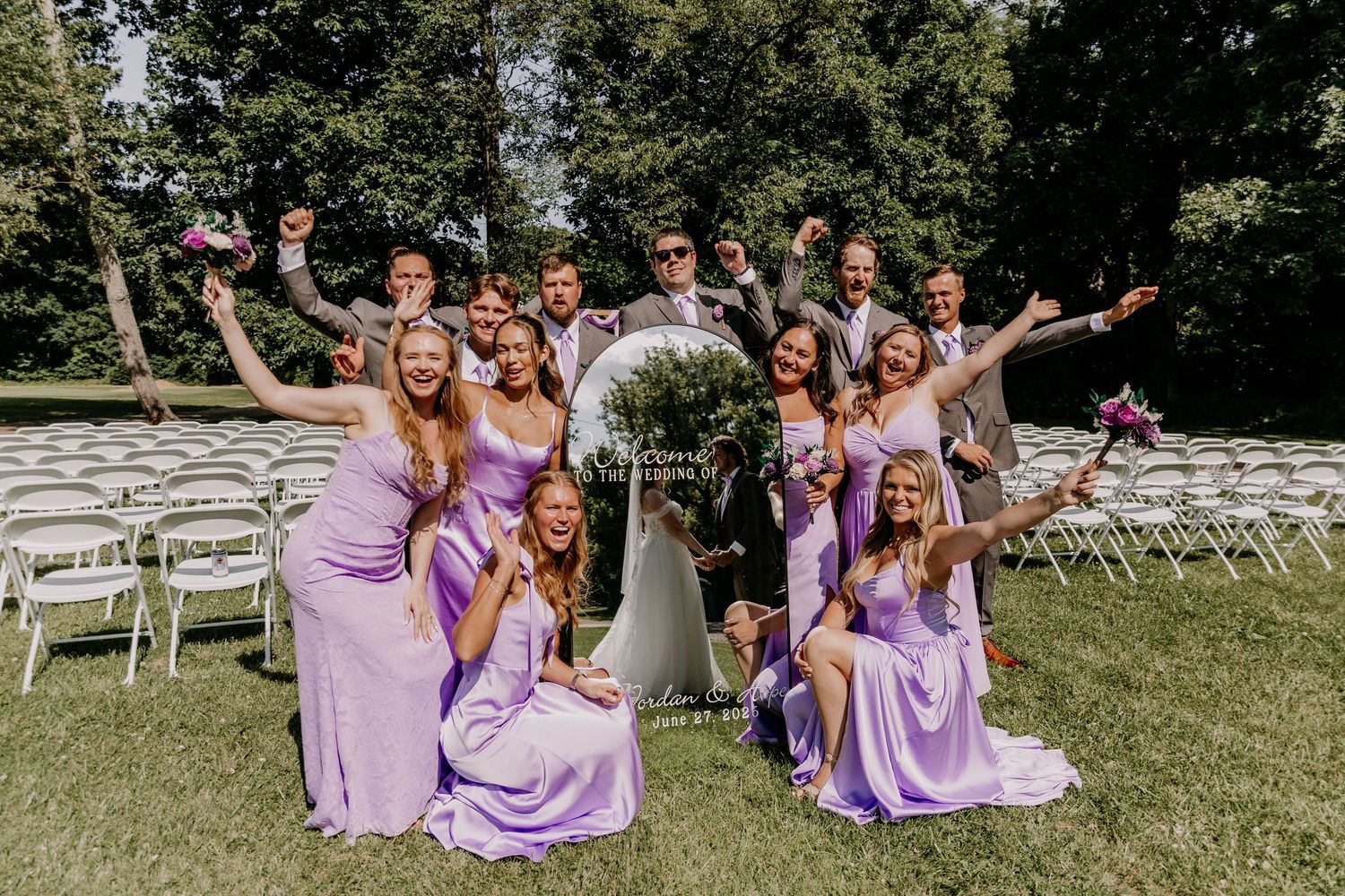Wedding party in purple bridesmaid dresses celebrating outdoors with groomsmen, cheering joyfully in a garden ceremony setting.