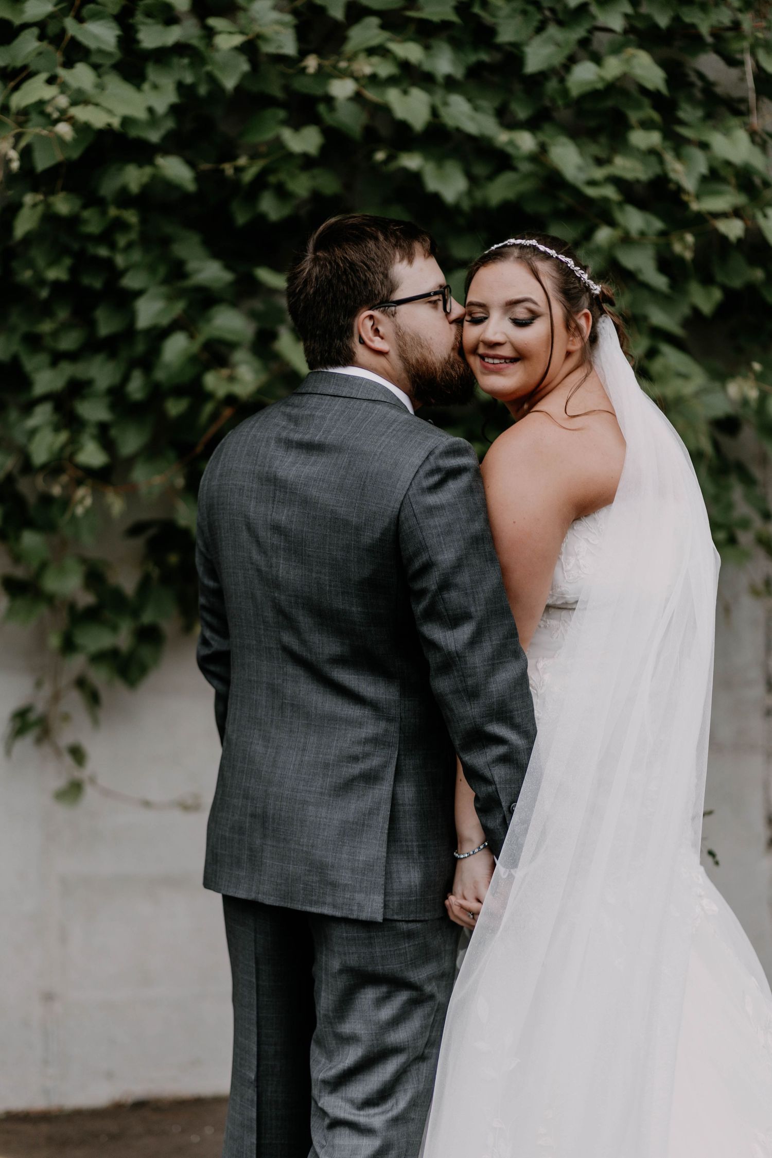 Intimate moment between newlyweds as groom in grey suit whispers to bride against ivy-covered wall.