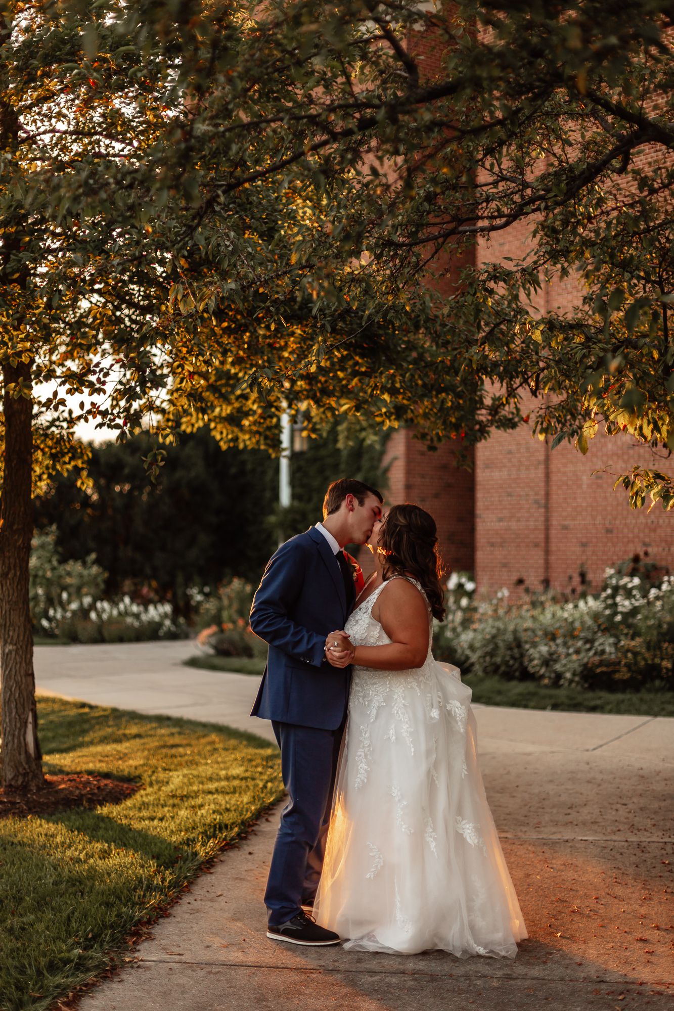 Romantic wedding couple embraces at sunset beneath autumn trees on brick path outside building.