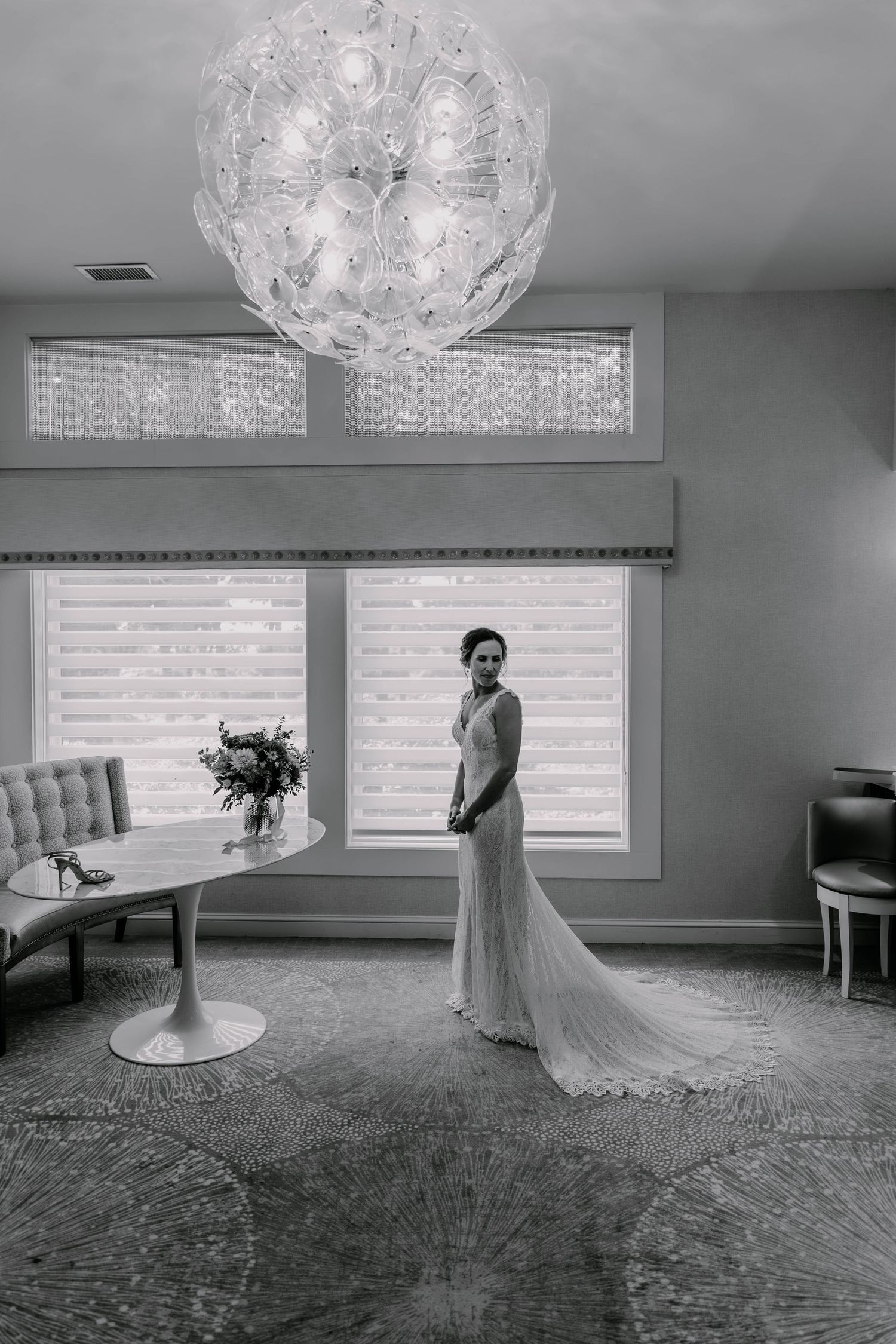 A black and white portrait of a bride in a flowing gown beneath an elegant crystal chandelier.