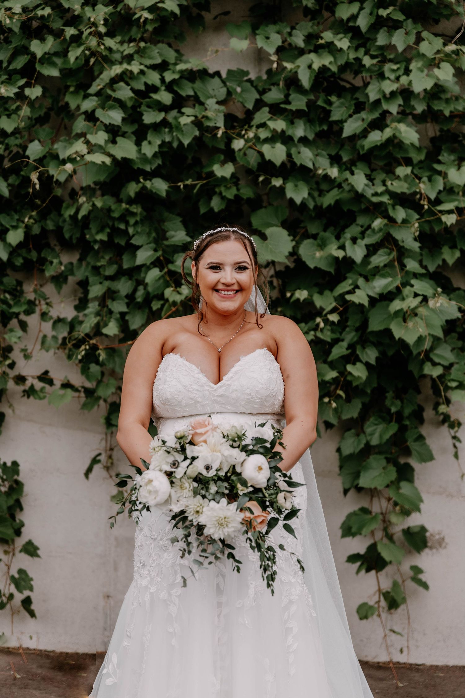 Bride in lace wedding gown poses with white floral bouquet against ivy wall.