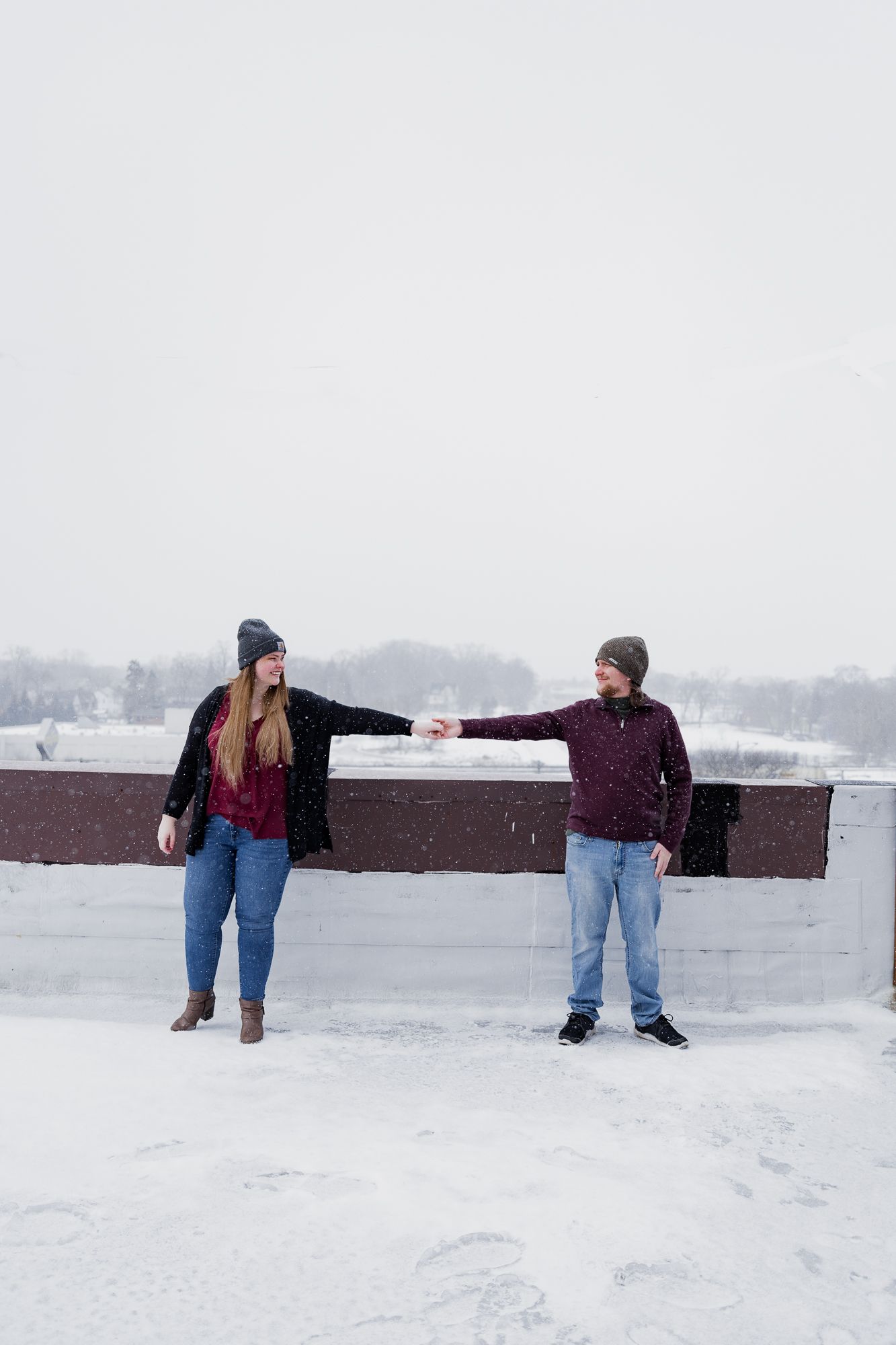 Two people playfully walking through a snowy landscape near a brick wall.