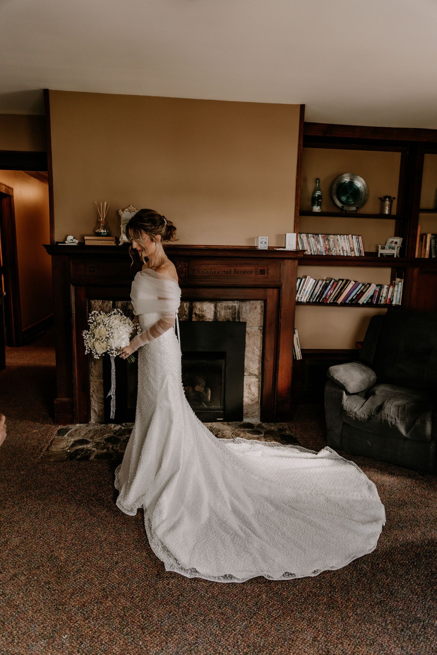 A bride in a flowing white wedding dress with a long train twirls gracefully in a rustic room with stone fireplace.