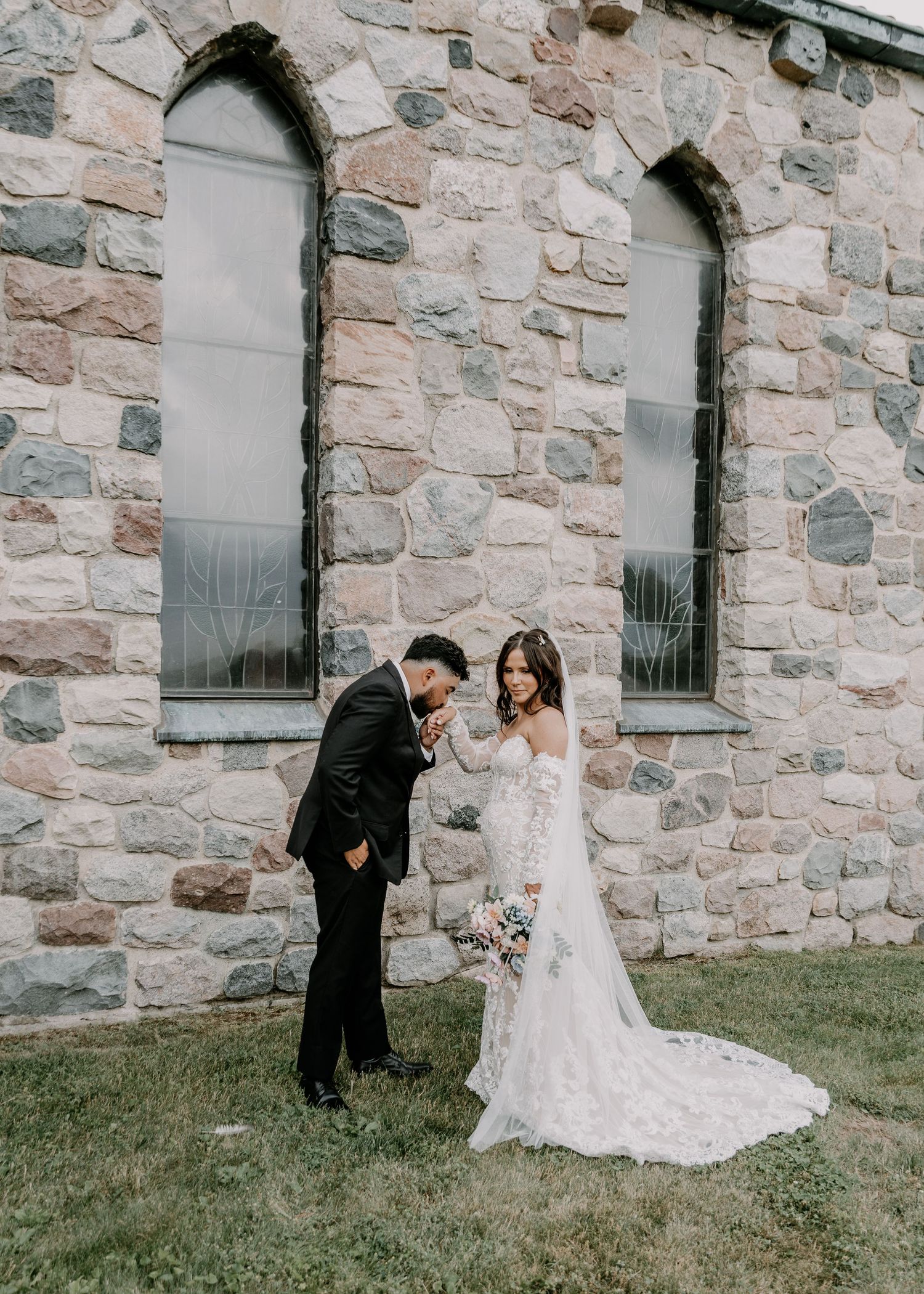 A couple shares a tender moment outside a stone church building with arched windows.