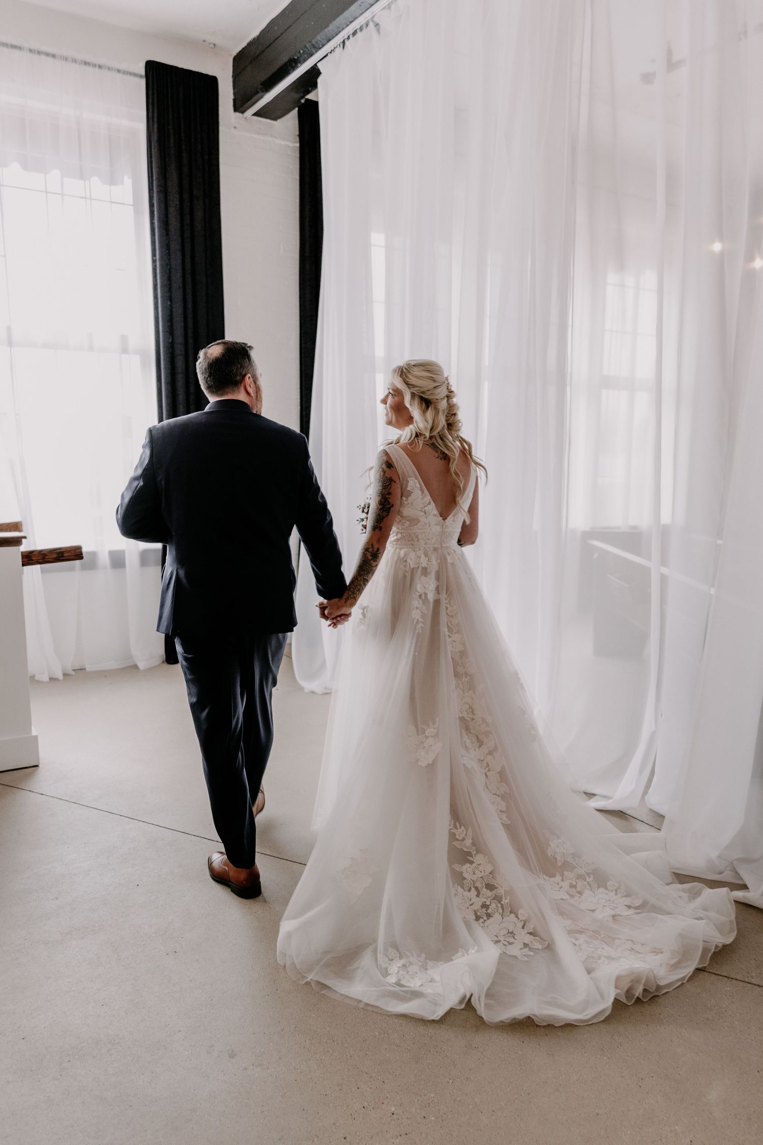 A couple walks through a bright white room with flowing curtains during their wedding day.