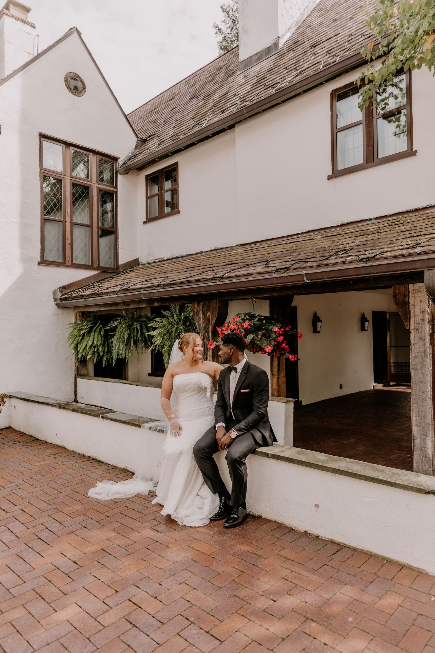 Bride in a strapless white gown and groom in a dark suit sit together on a low wall outside a white venue with a covered brick patio.