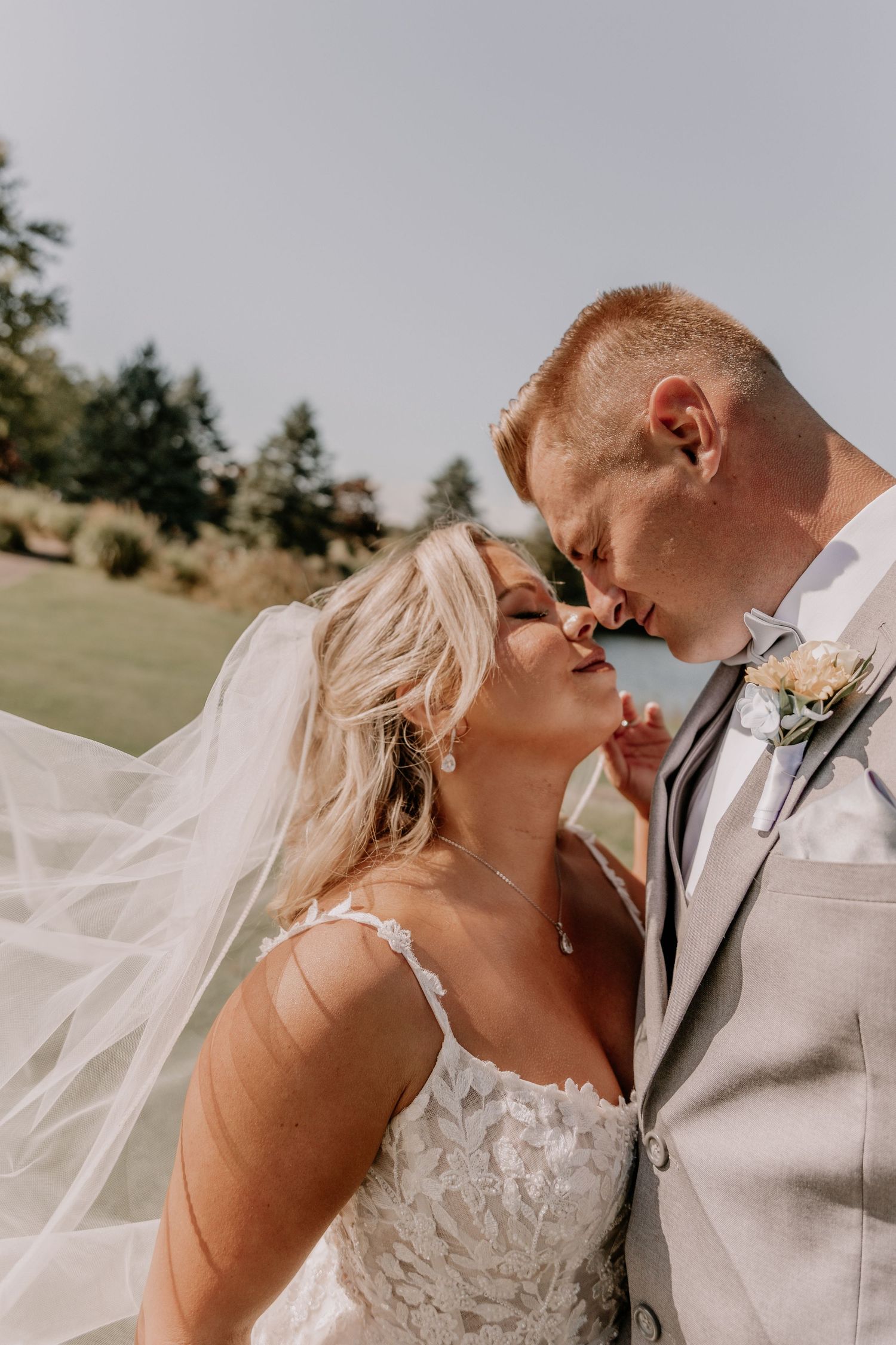 A bride and groom share a romantic kiss outdoors, her veil flowing in the breeze, wearing a lace gown and gray suit.