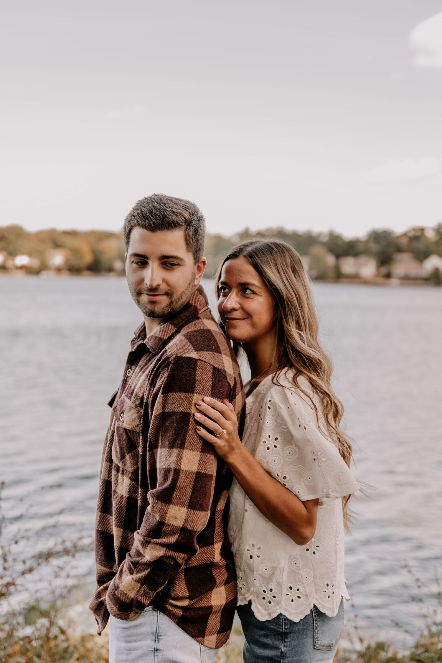 Couple posing by a lake, woman in white top rests hands on man in plaid shirt, fall foliage backdrop.