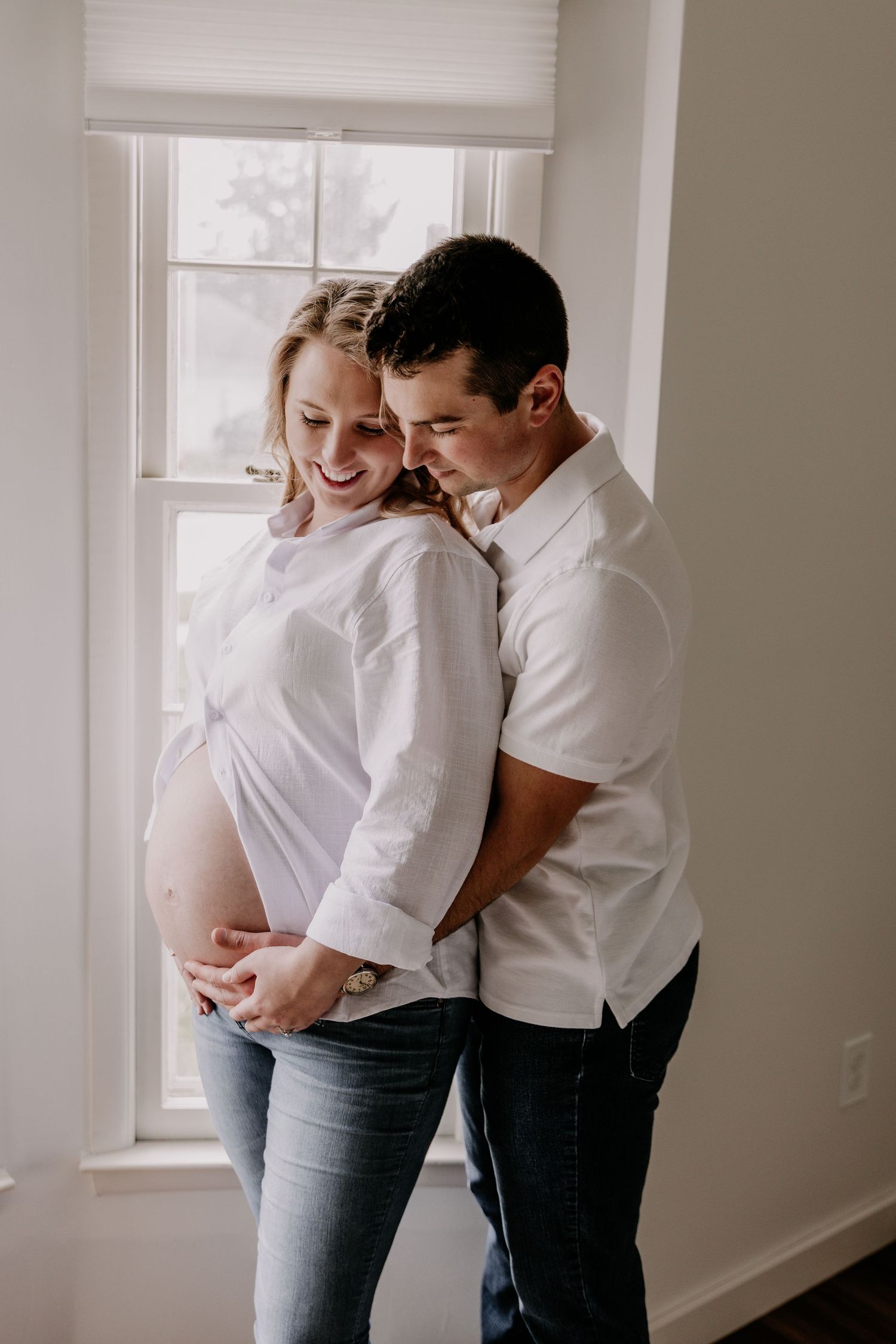 Pregnant couple embracing near window, woman in white shirt showing baby bump, man hugging her from behind.