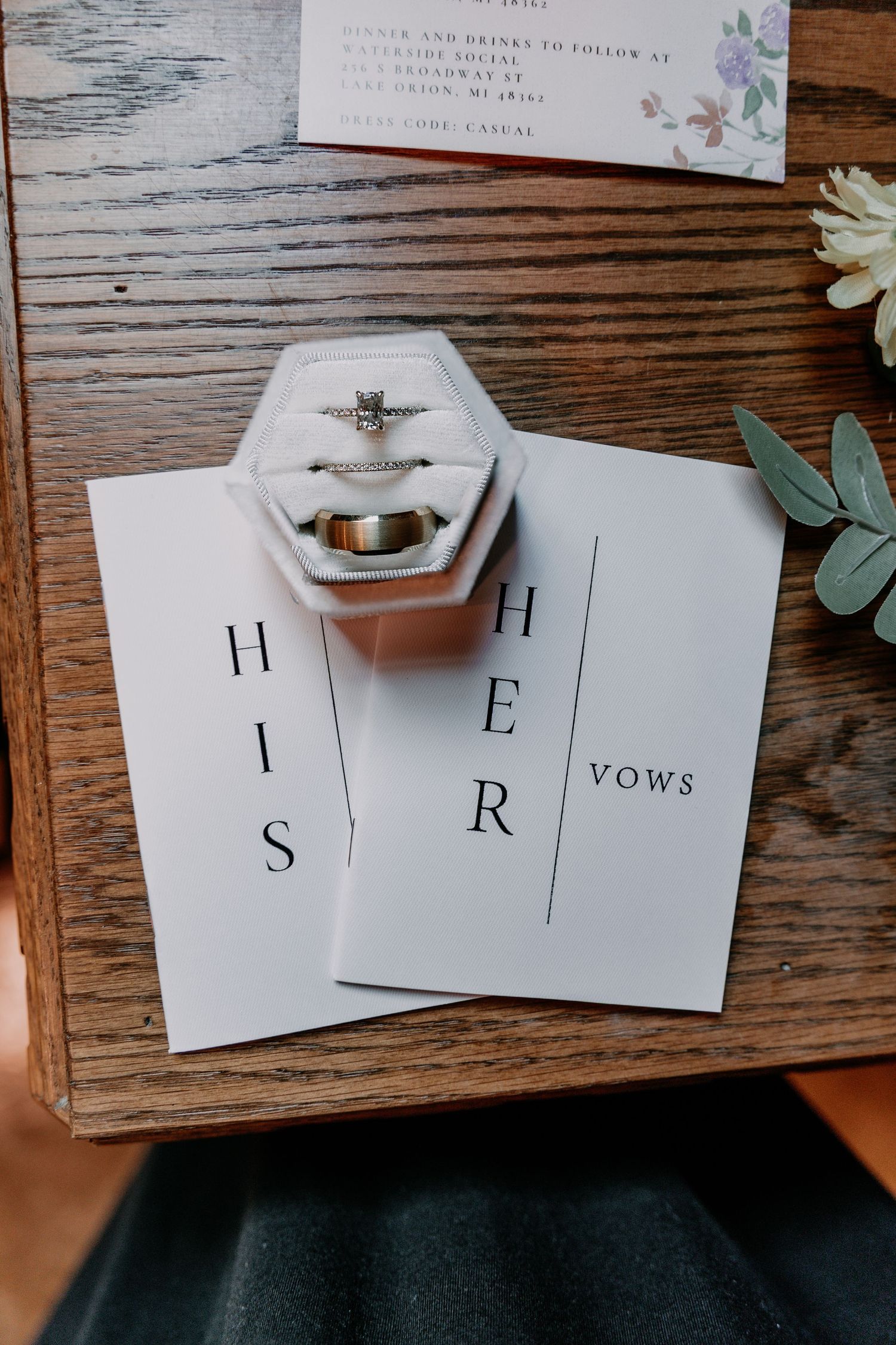 Wedding vow book pages spread across a rustic wooden table with scattered rose petals and greenery.