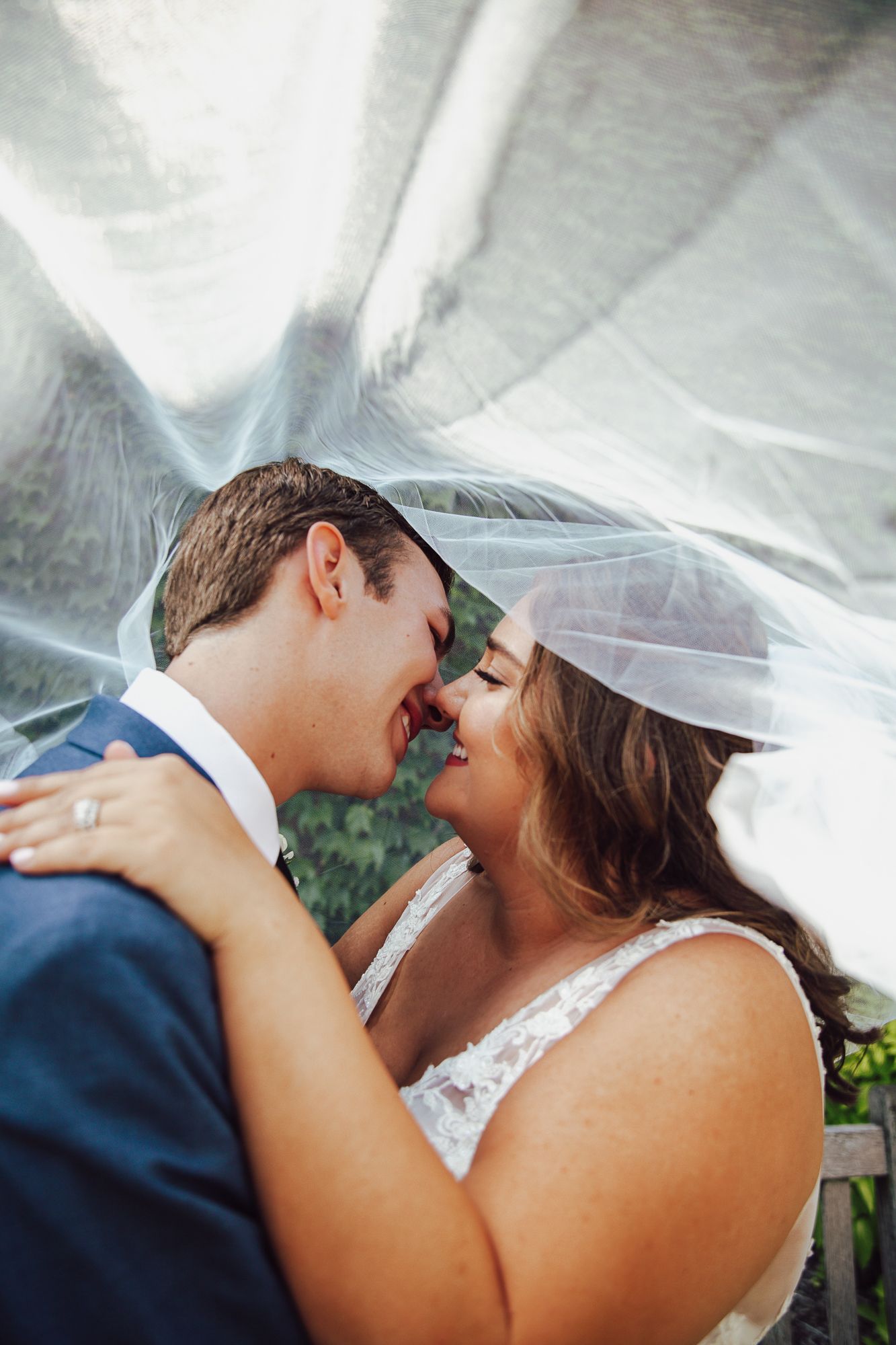 Romantic couple embraces under flowing wedding veil during outdoor garden ceremony.