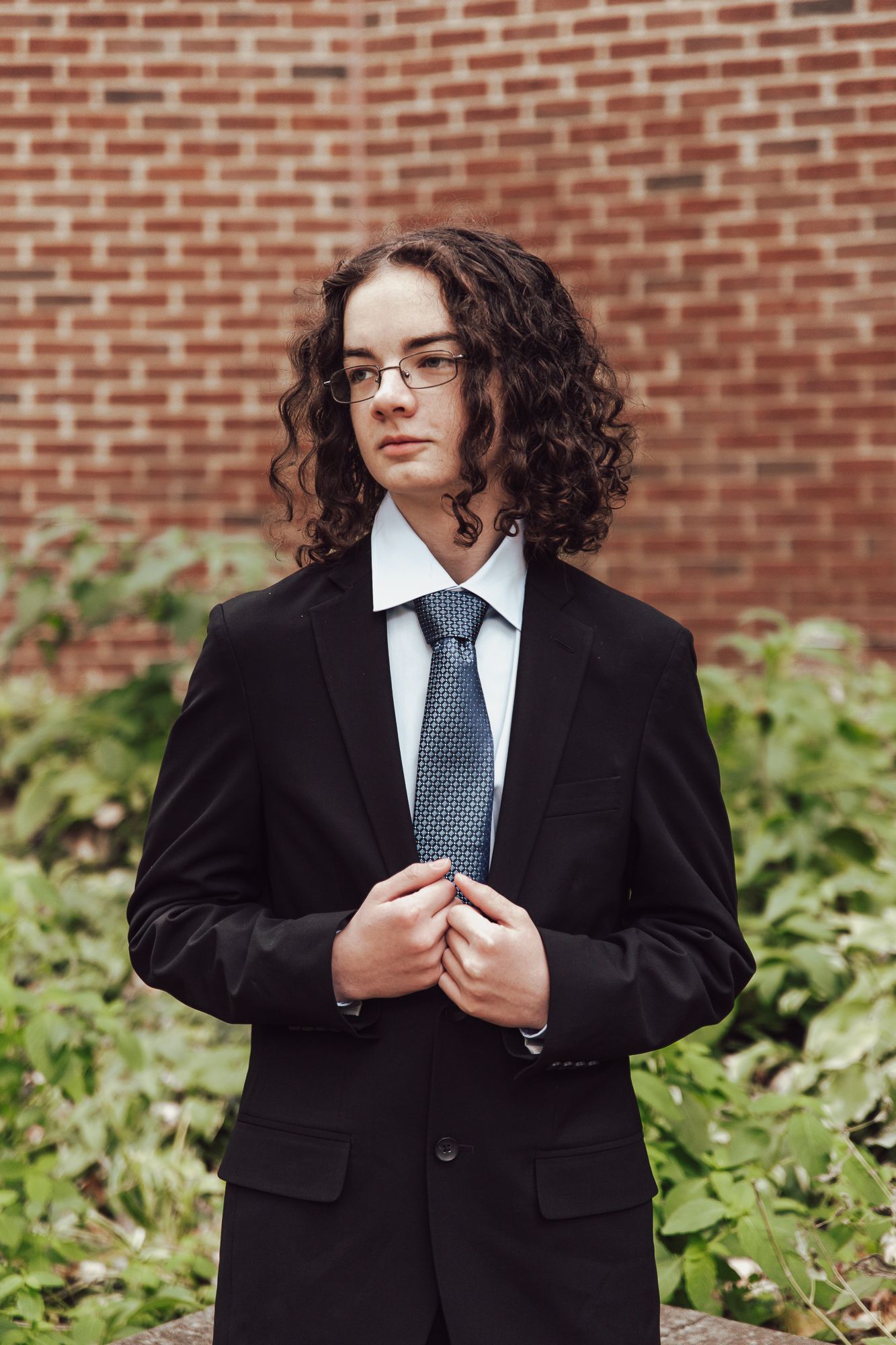 Portrait of a person in a formal black suit and tie with curly hair against a brick wall backdrop.
