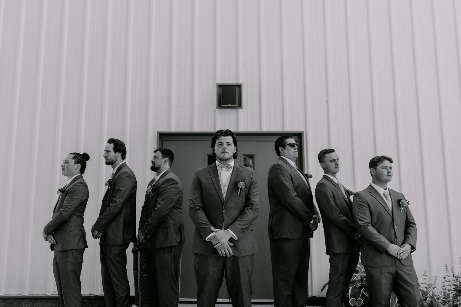 Black and white photo of groom and groomsmen in suits posing confidently outside a modern barn wedding venue.