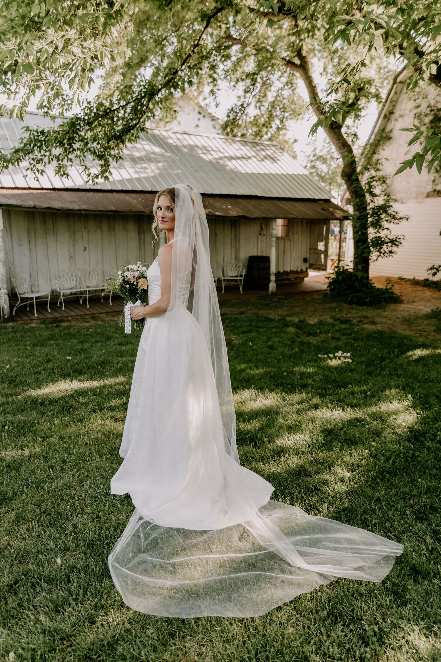 Bride in flowing white gown and long veil holding bouquet stands on green lawn near rustic barn.