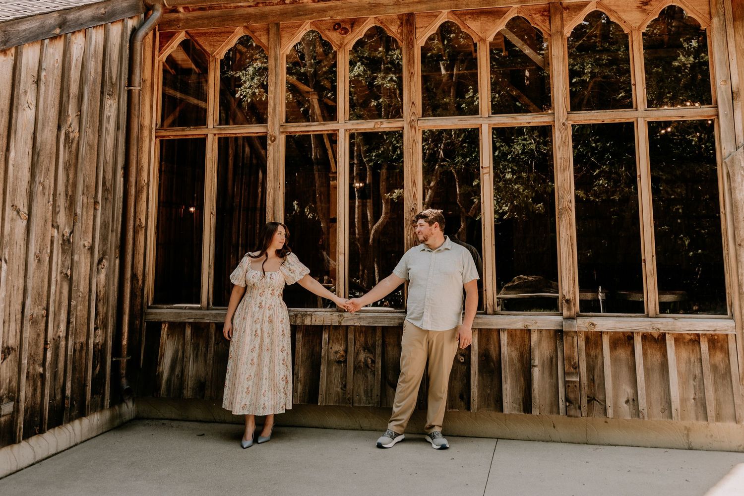 Couple holding hands in front of rustic wooden building with arched windows during outdoor engagement photo session.