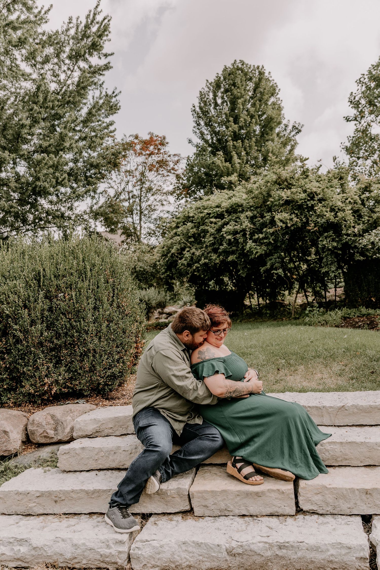 A couple shares a tender moment sitting on stone steps surrounded by lush greenery, the woman in a green maternity dress.