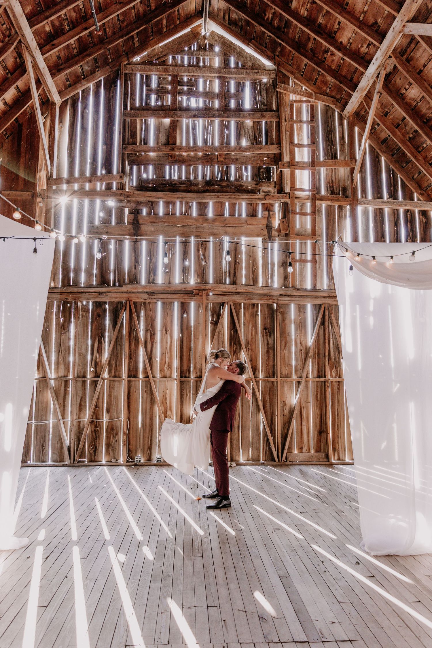 A rustic barn interior with wooden beams, white drapes, and string lights creating a romantic wedding venue atmosphere.