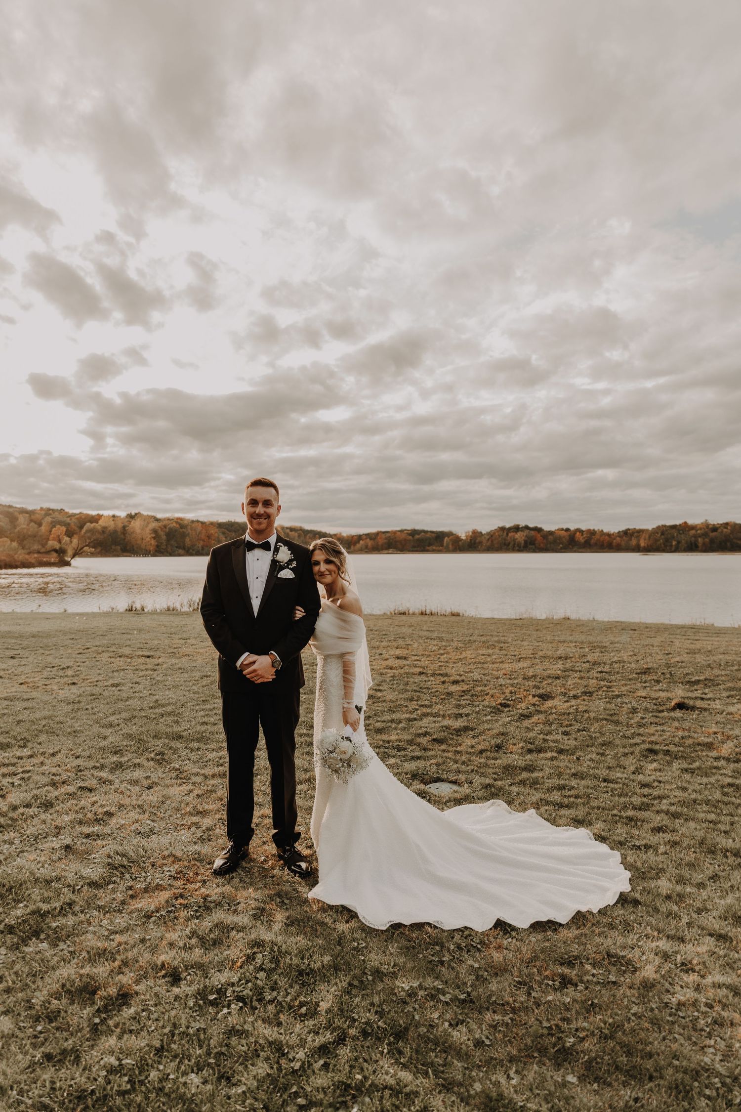 A couple poses for wedding photos on a grassy lakeside under dramatic cloudy skies during autumn.