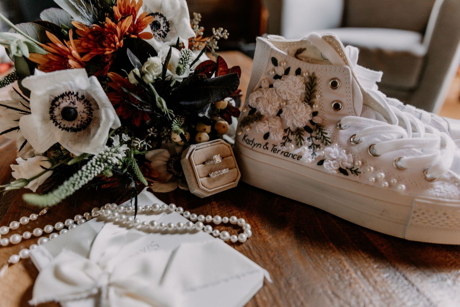 Bridal accessories including white shoes, pearl necklaces, rings, and floral decor arranged on a rustic wooden surface.