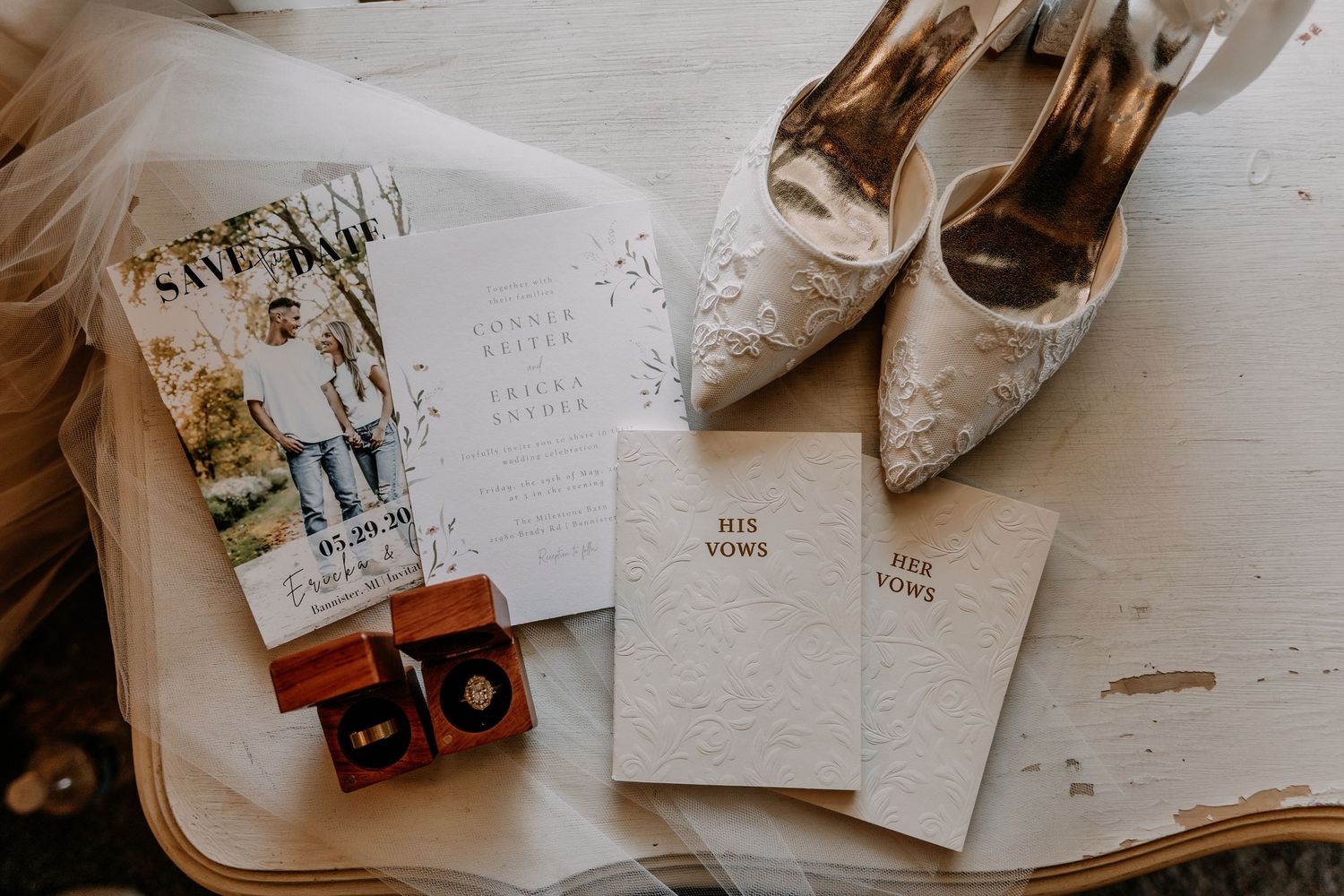 Wedding flat lay featuring save the date cards, vow booklets, gold rings, and lace bridal heels on a vintage tray.