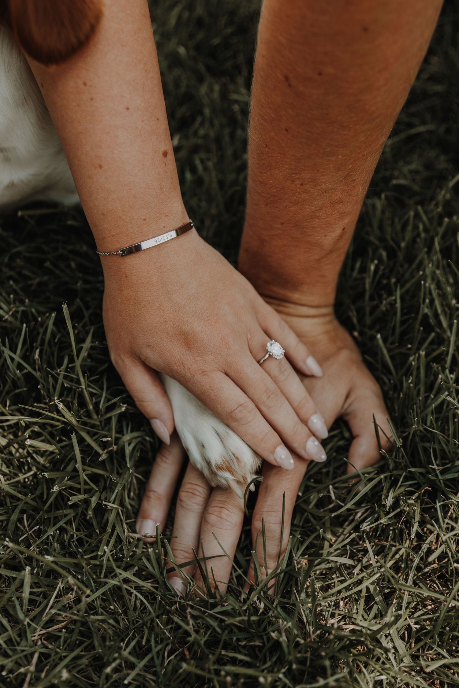 Close-up of hands wearing engagement rings resting on grass, showcasing white nail polish and delicate jewelry.