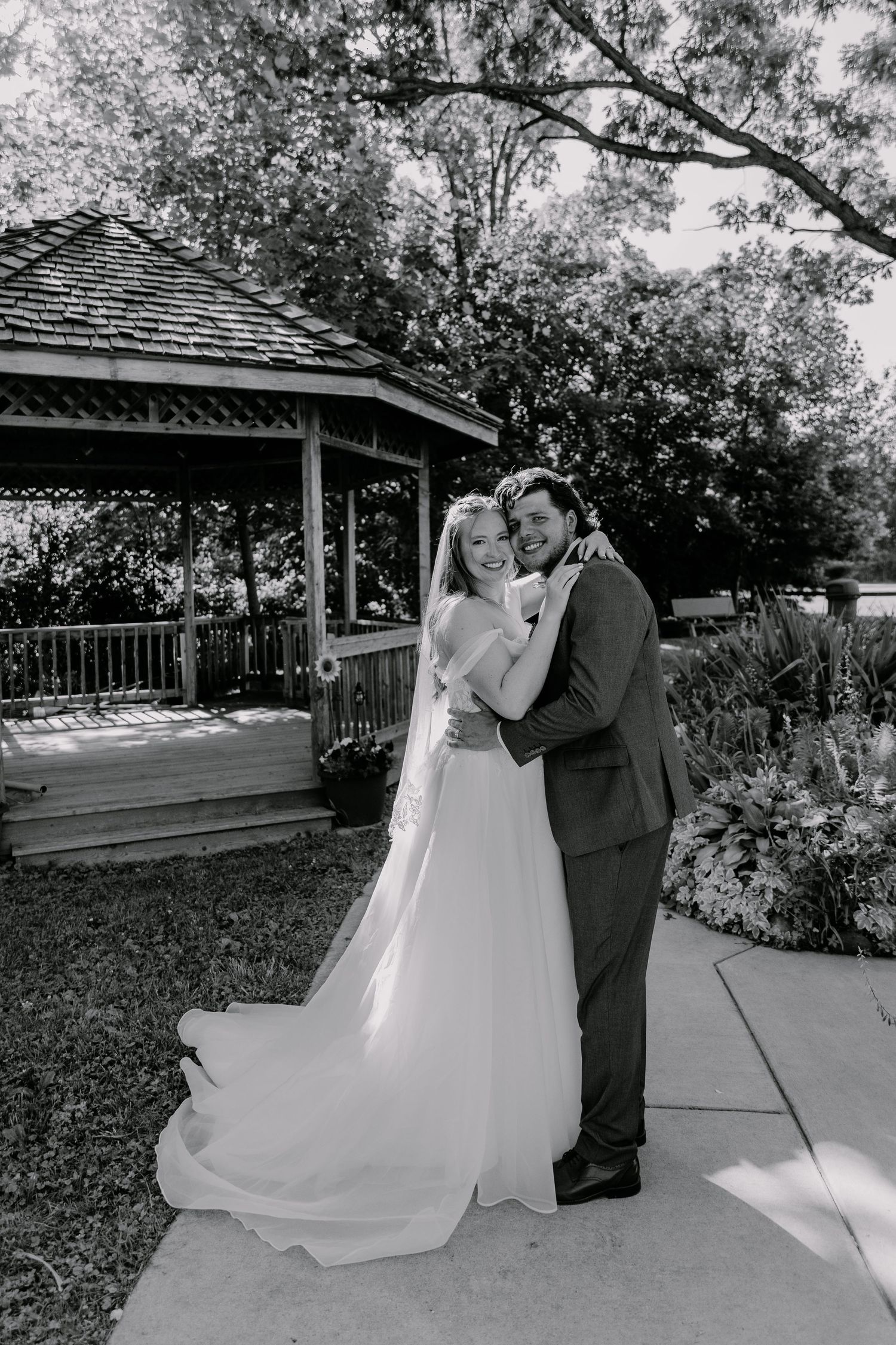 Black and white photo of a bride and groom embracing near a gazebo surrounded by lush trees and gardens.