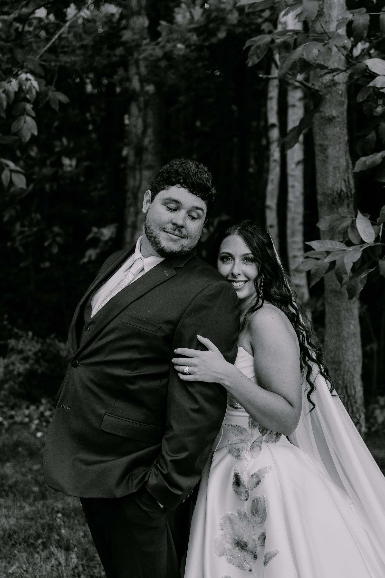 Black and white wedding photo of smiling bride in floral gown embracing groom in dark suit outdoors.