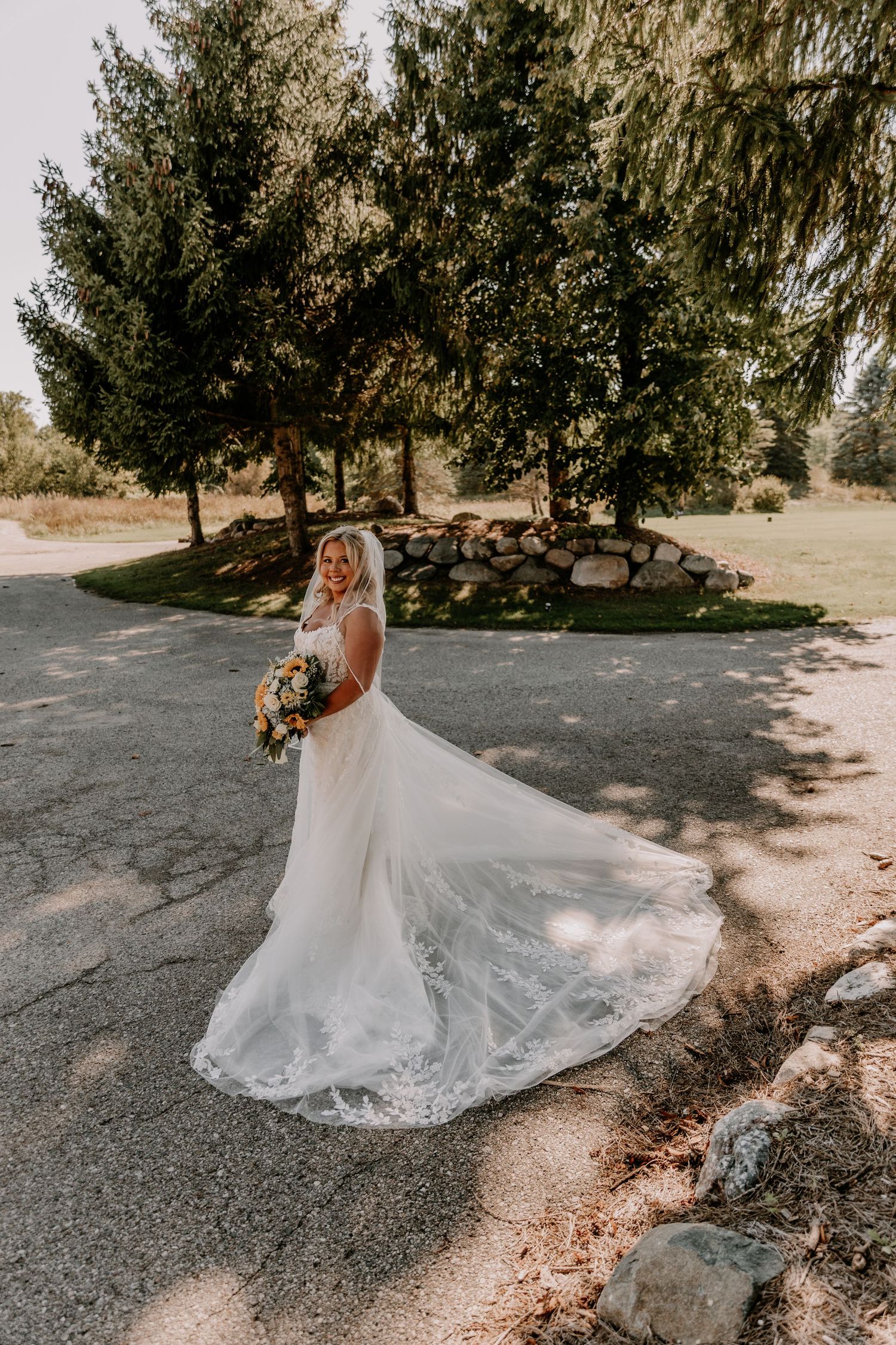 Bride in a flowing white lace wedding gown holding a bouquet, posing outdoors near trees on a sunny day.