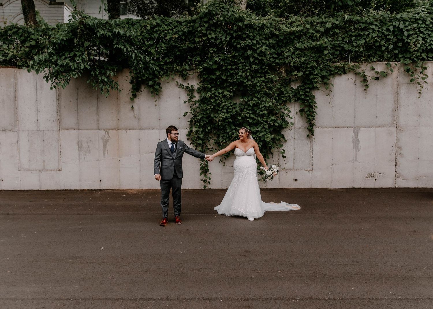 Full-length wedding portrait with dramatic long veil against concrete and ivy wall.