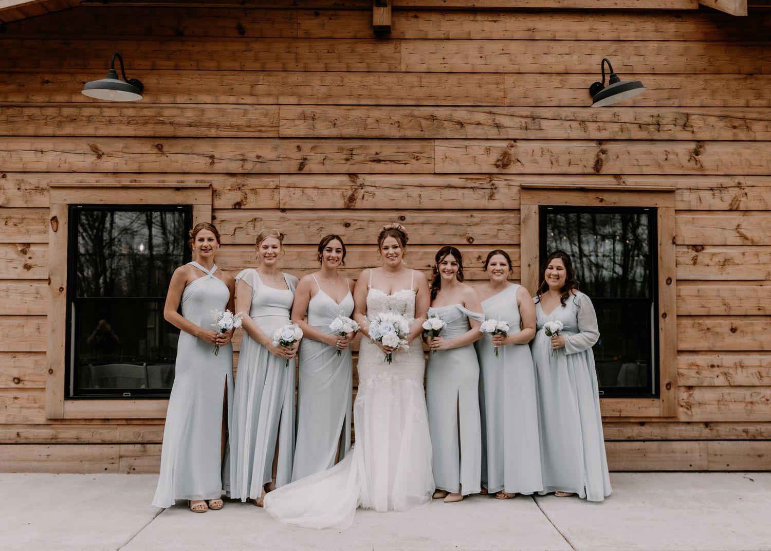 Bridesmaids in light gray dresses pose with bride against wooden barn exterior.