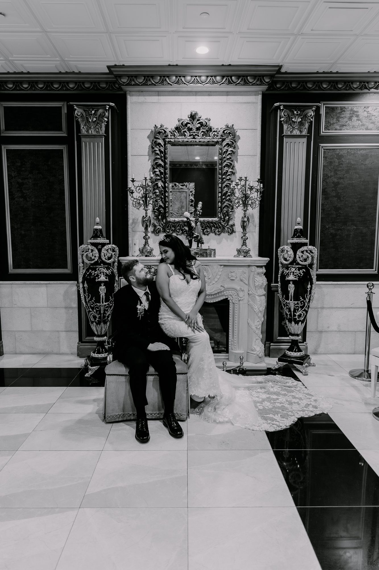 A newlywed couple sits together in an elegant room with ornate decor and a decorative fireplace mantel.