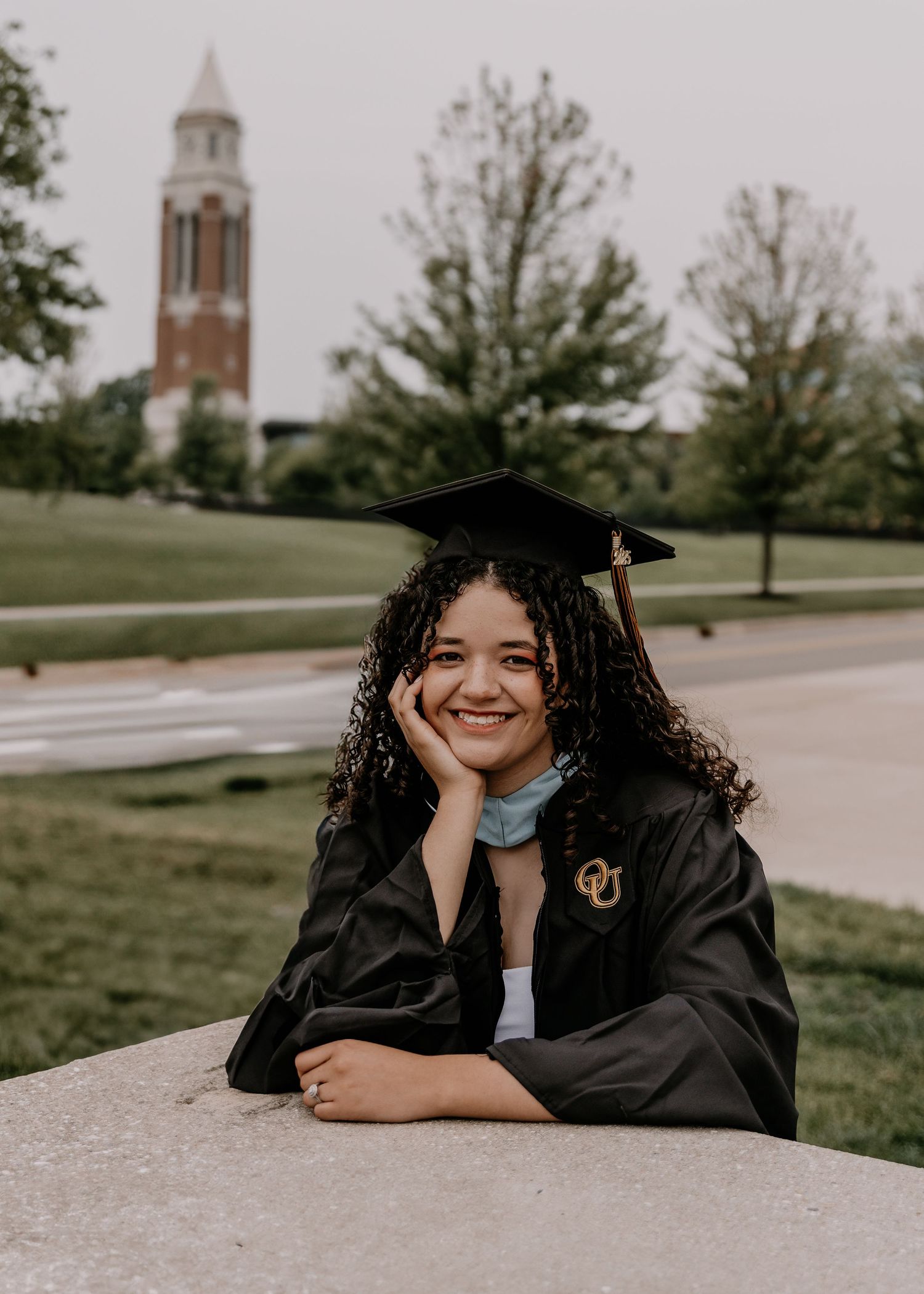 Graduate wearing black cap and gown poses on campus with bell tower and green lawn visible in background.