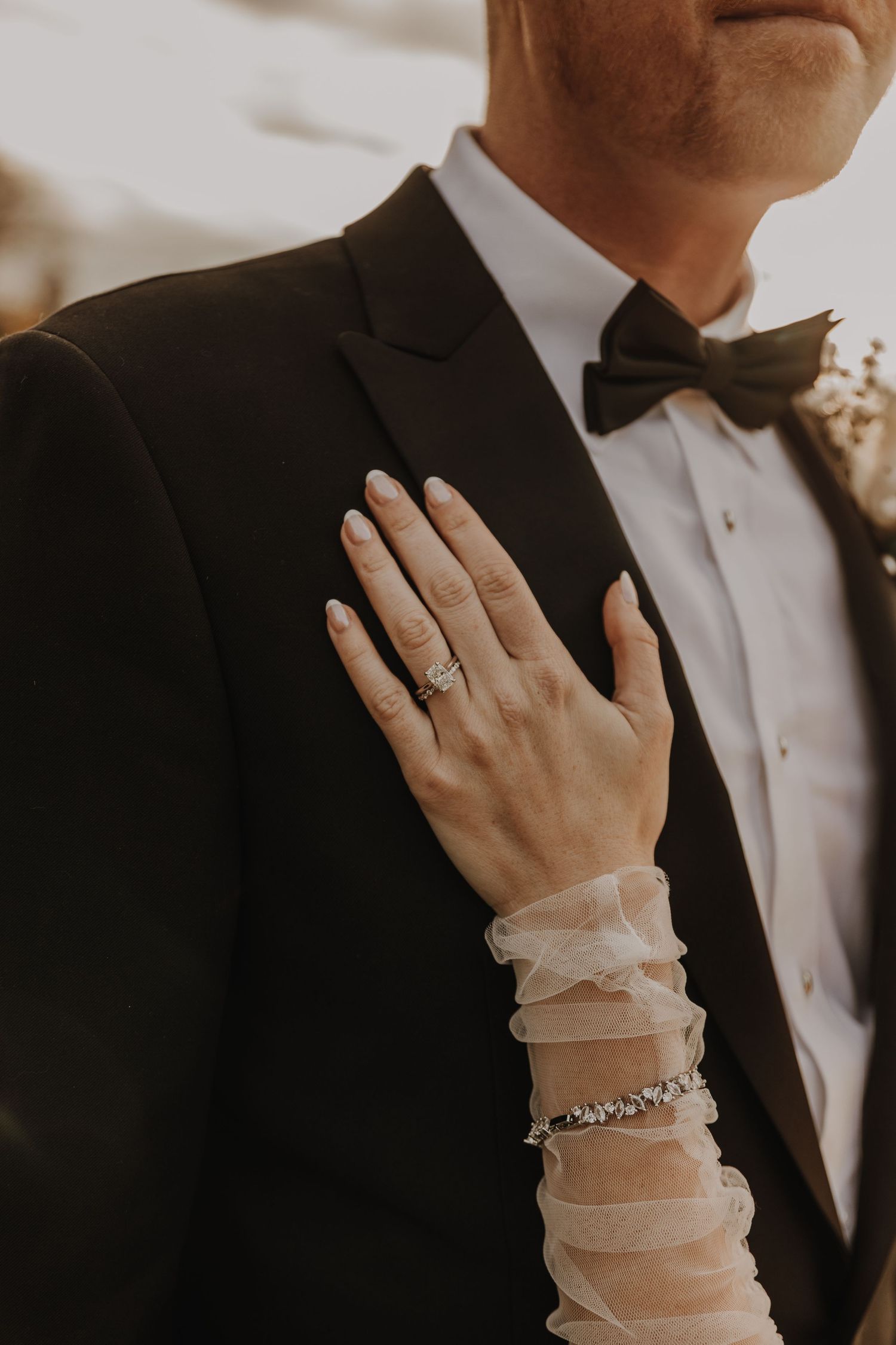Close up of bride's hand with engagement ring resting on groom's formal black tuxedo.