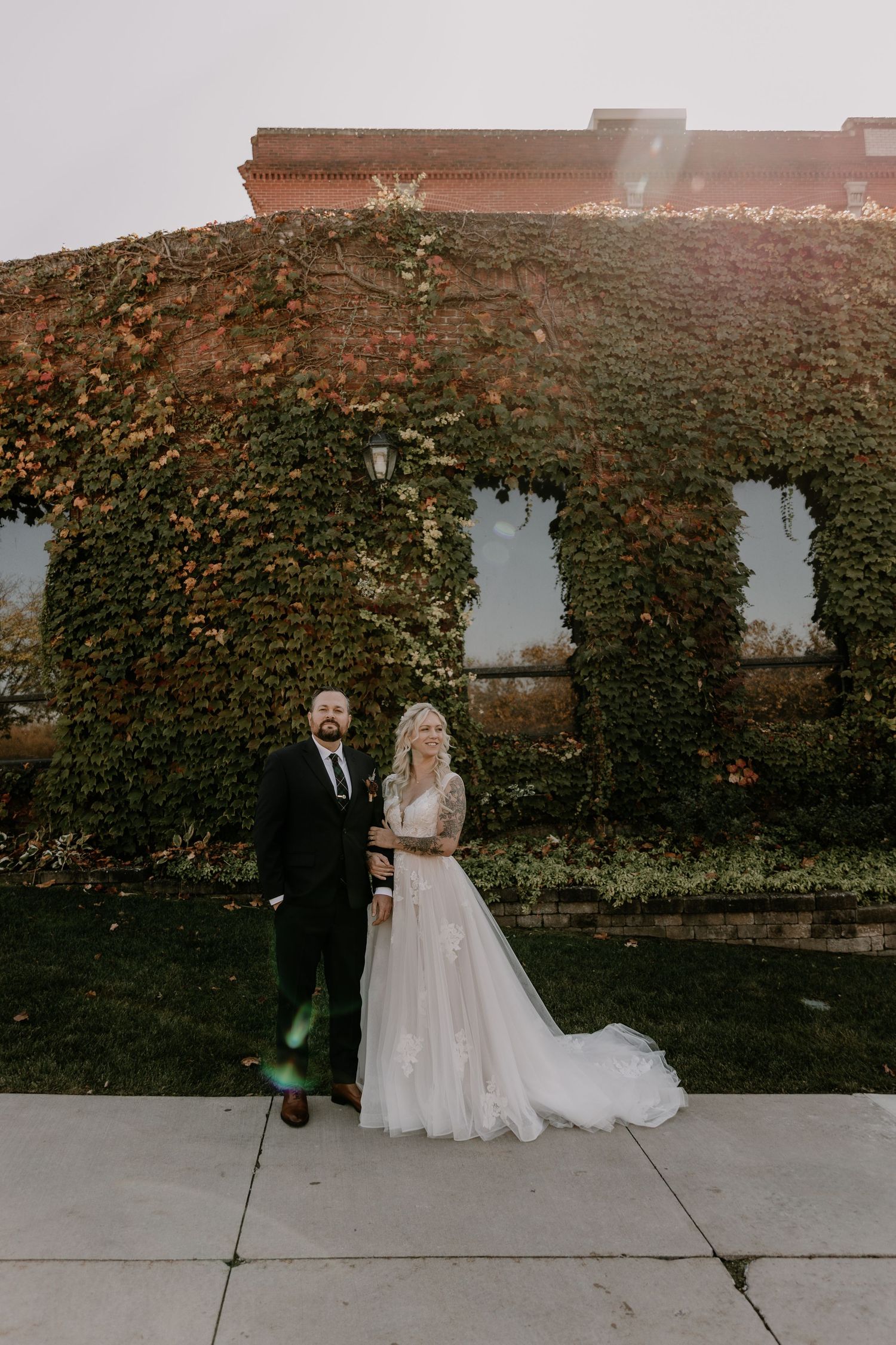 A couple poses in front of an ivy-covered brick wall on their wedding day, with a beautiful trailing white dress.