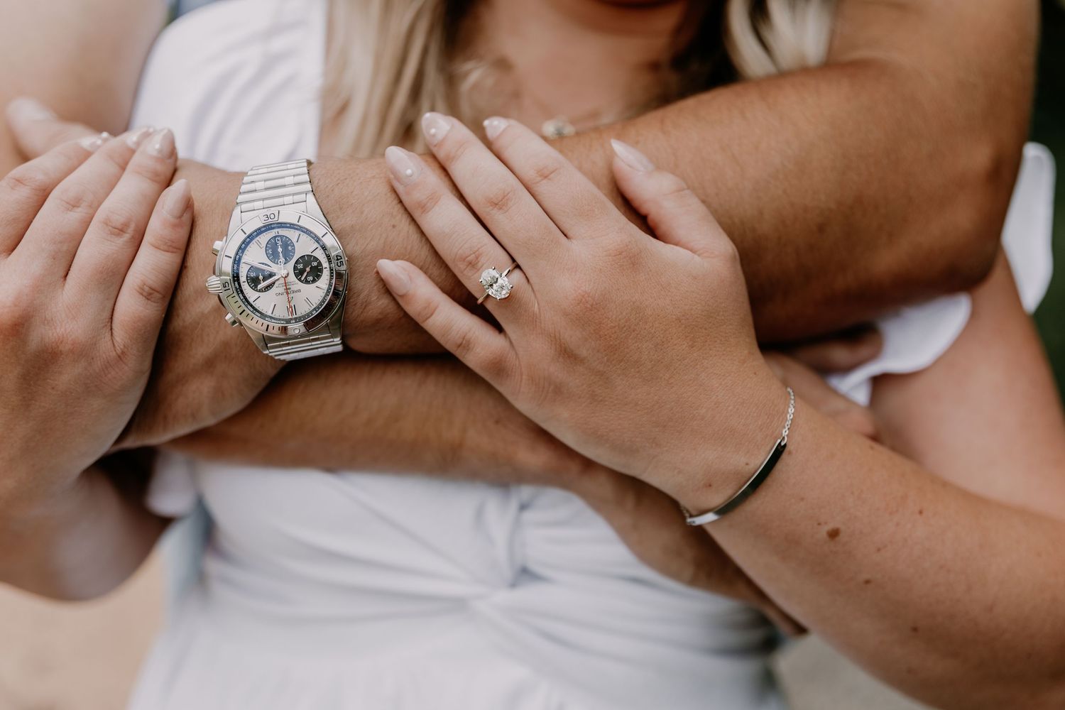 Two hands intertwined showing engagement ring and luxury chronograph watch against white clothing in intimate pose.