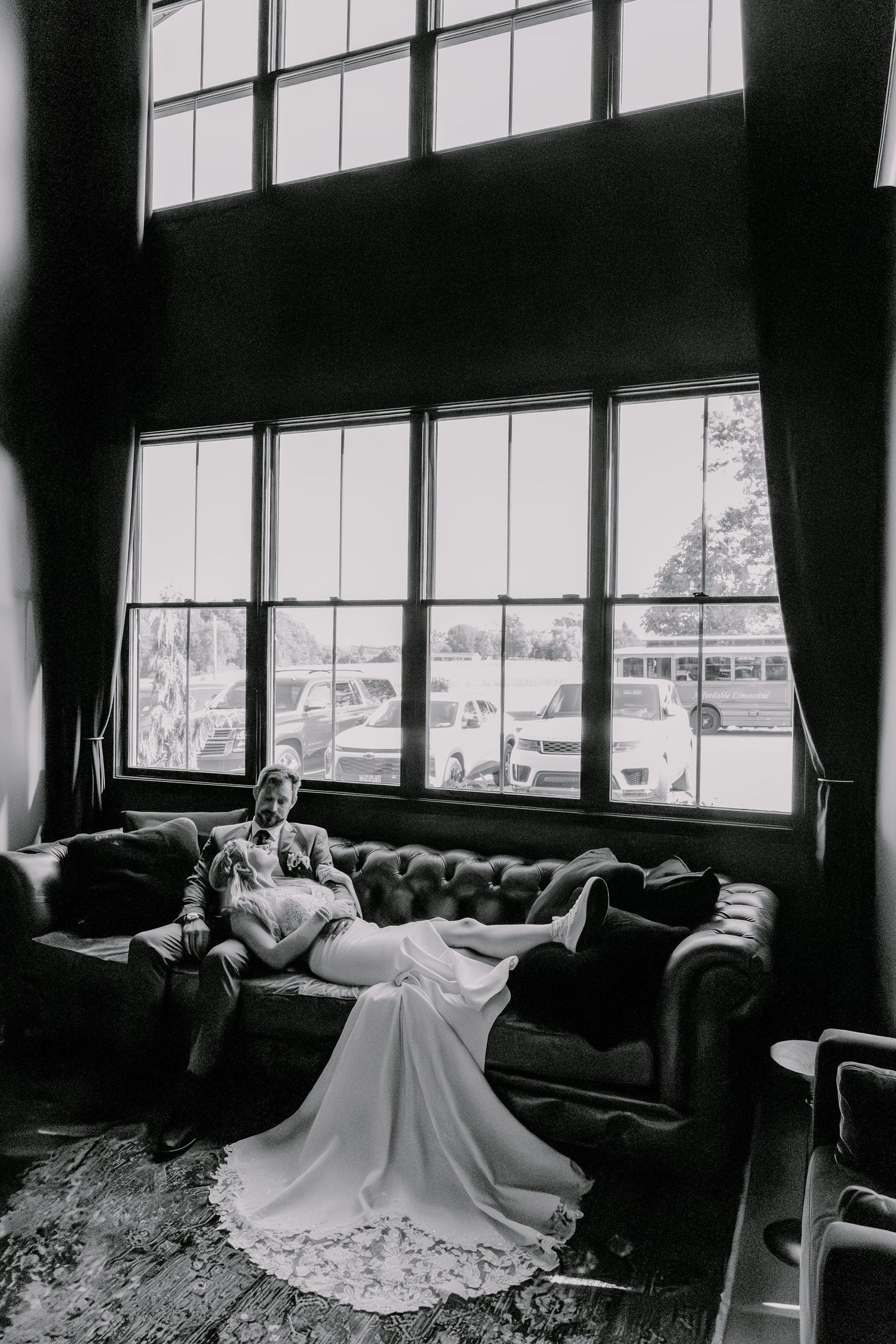 Elegant wedding dress spread across a vintage couch near large industrial windows in black and white.