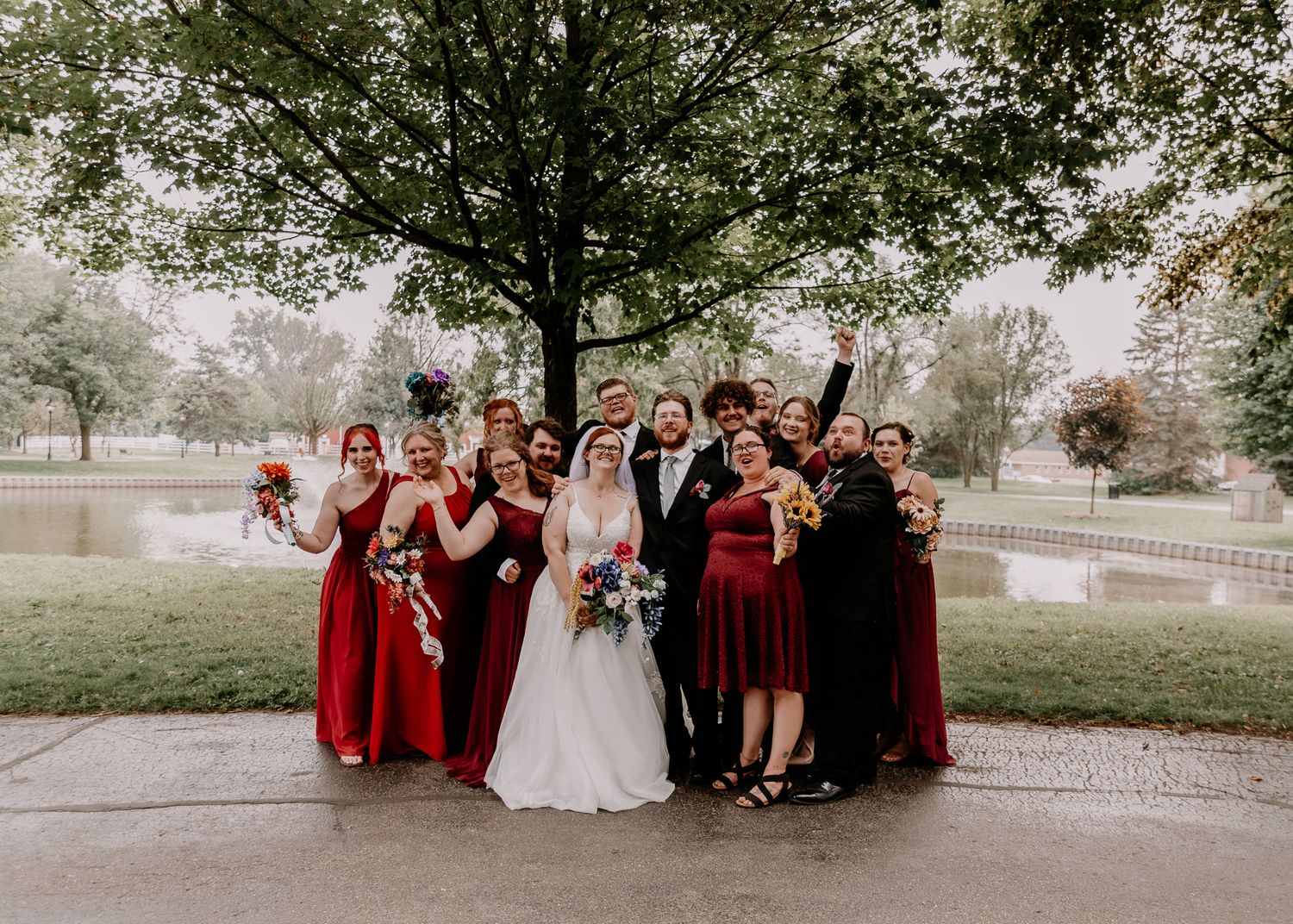 Wedding party in red dresses and formal attire poses together outdoors under trees.