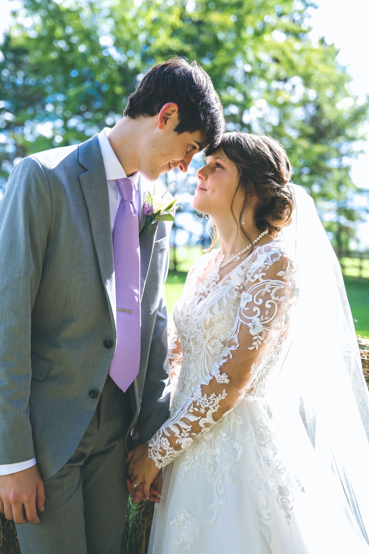 Wedding couple in grey suit and lace dress pose together in a sunlit garden setting.