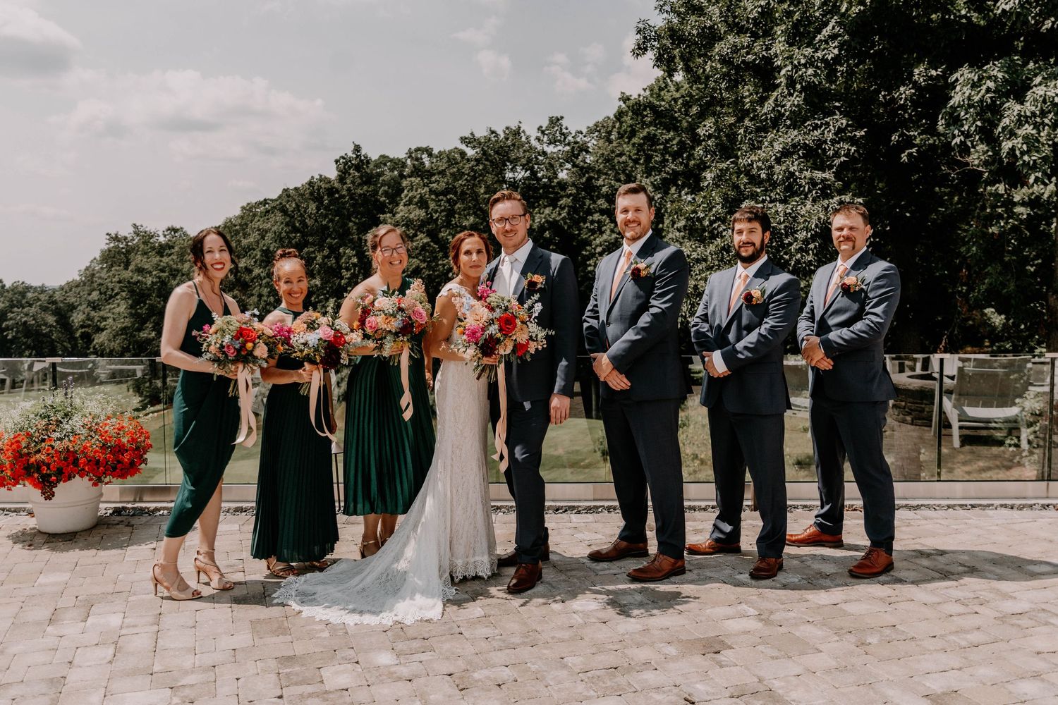 Wedding party in navy suits and dark dresses poses outdoors on stone patio with bright floral bouquets.