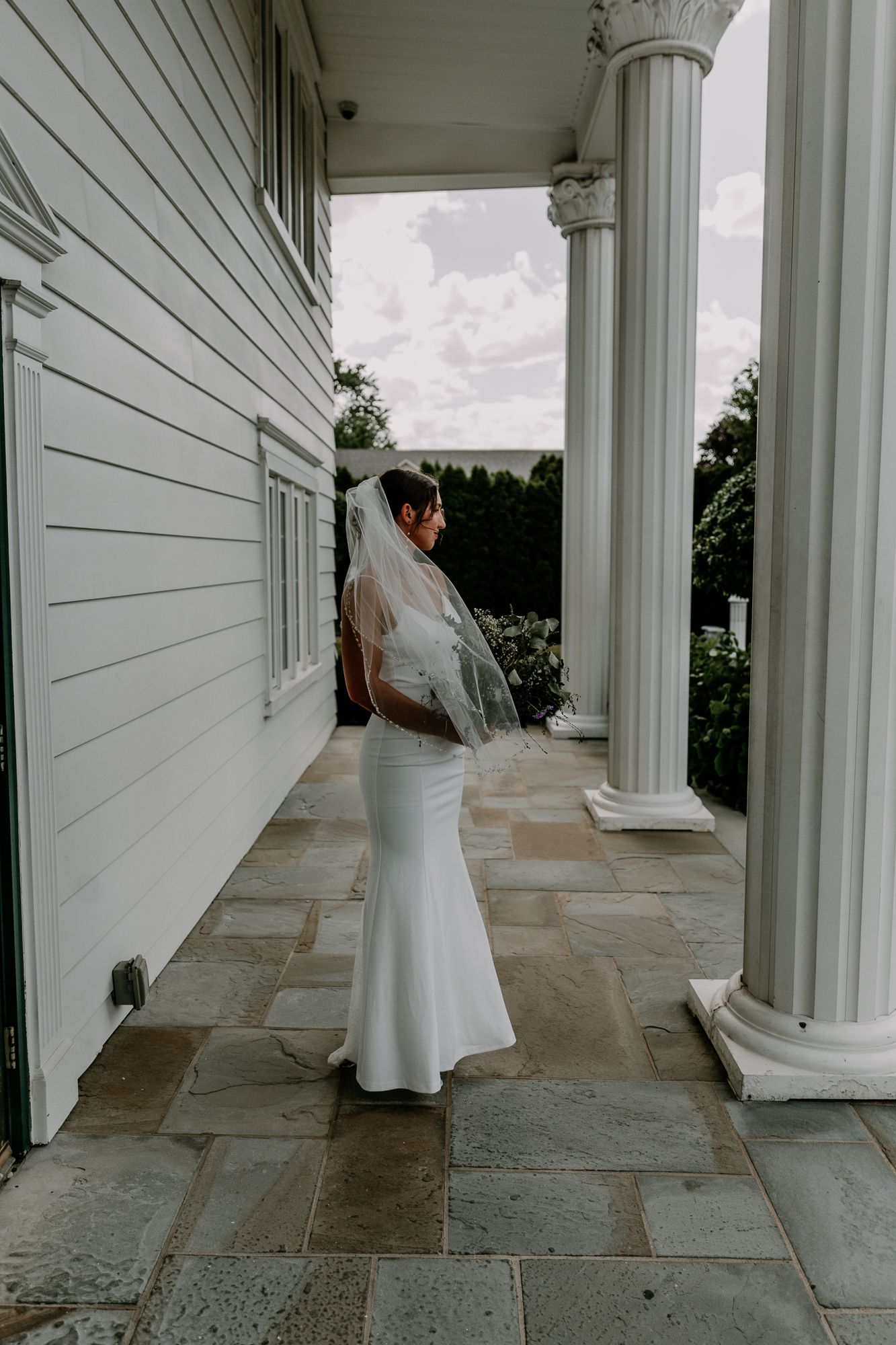 A bride in a sleek white gown poses on a stone porch with classical white columns.