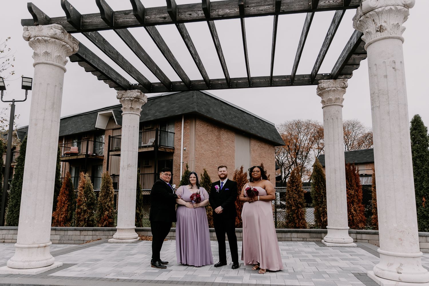 Wedding party stands under a white pergola with decorative columns during an outdoor ceremony.