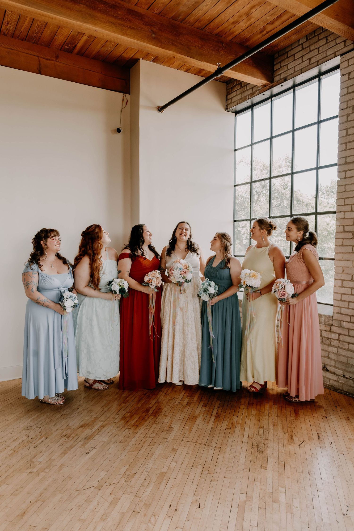Wedding party in mix-and-match pastel and jewel-toned dresses standing together in industrial loft space.