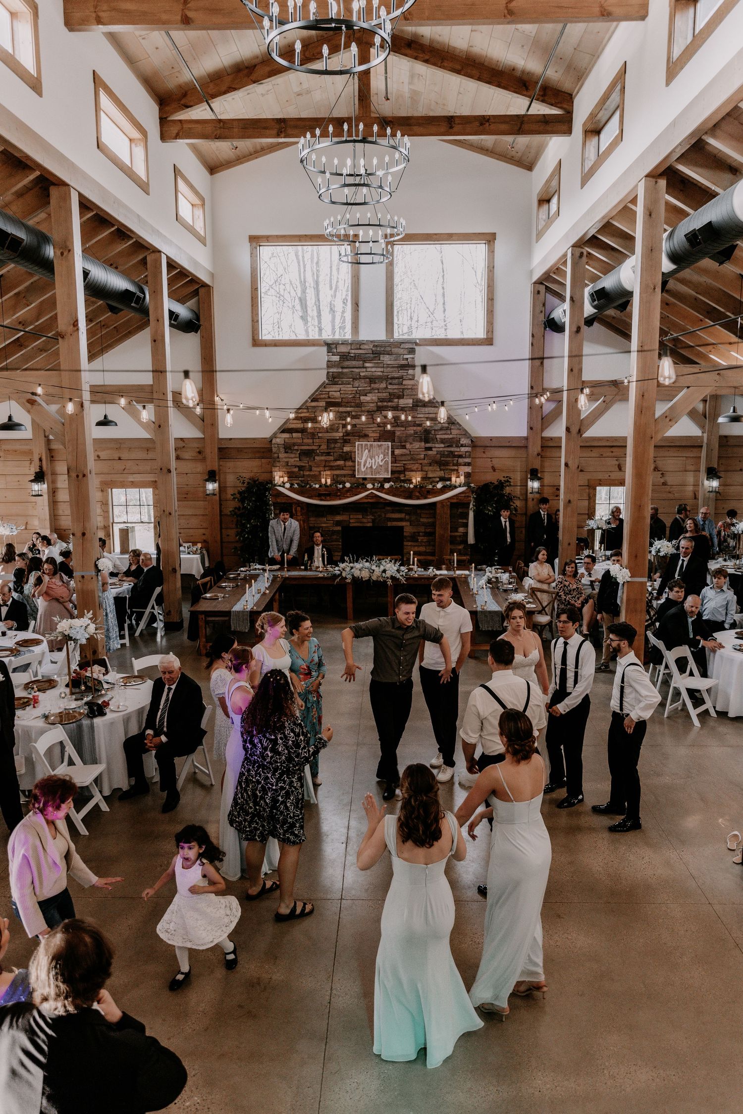 Wedding reception guests dancing in rustic barn venue with elegant chandeliers and stone fireplace.