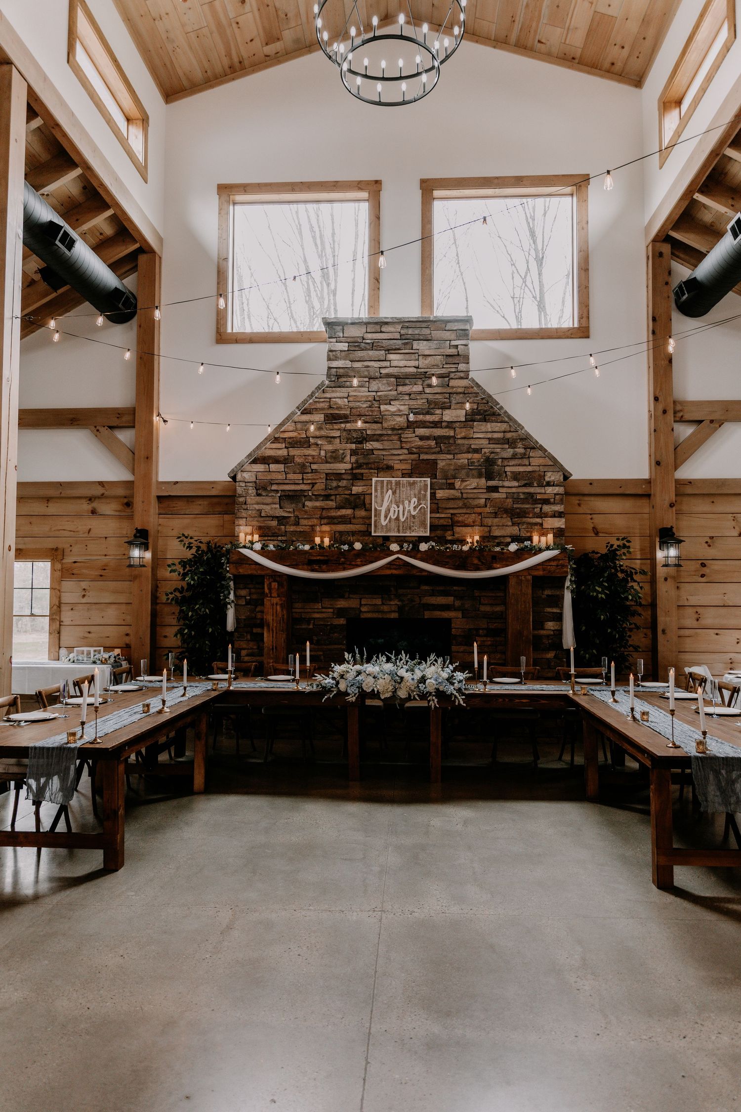 Rustic barn wedding venue interior with stone fireplace, wooden beams, and string lights.