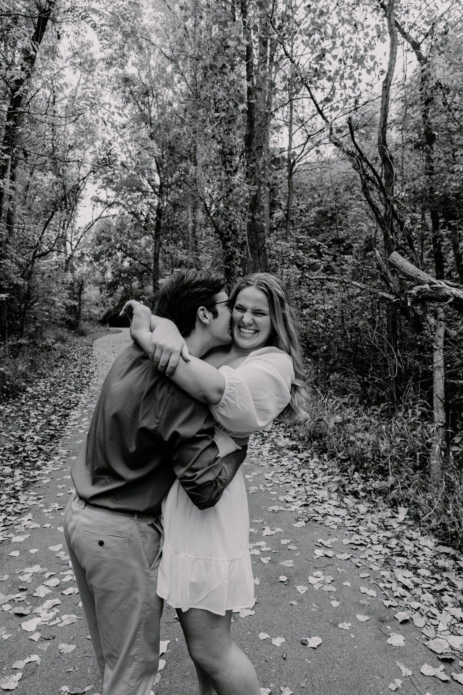 A couple shares a playful embrace on a forest path covered in fallen leaves.