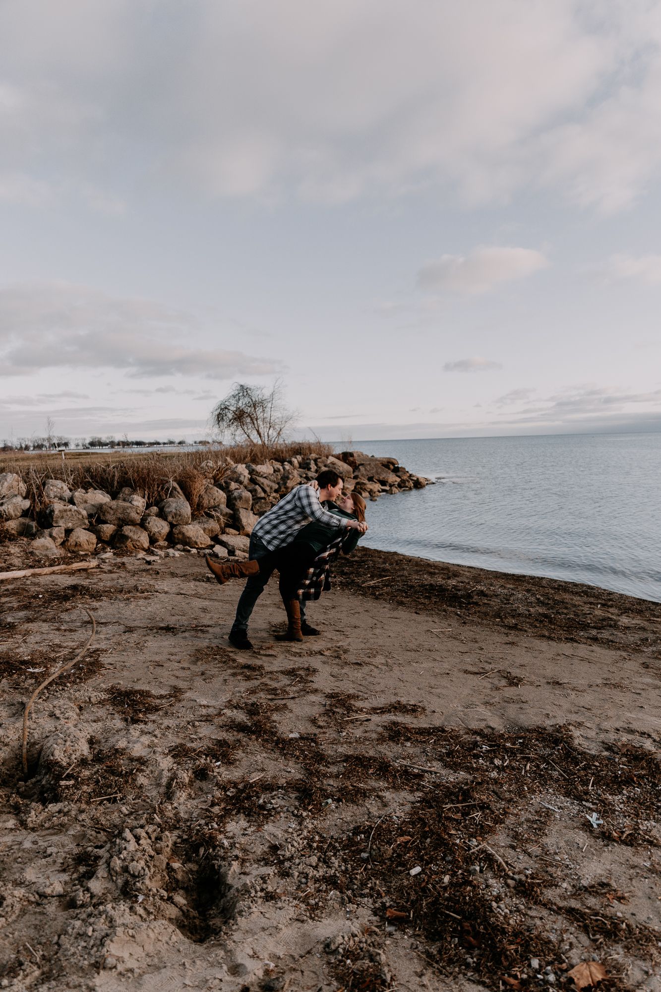 Couple shares intimate moment on rocky shoreline with lake and cloudy sky in background.