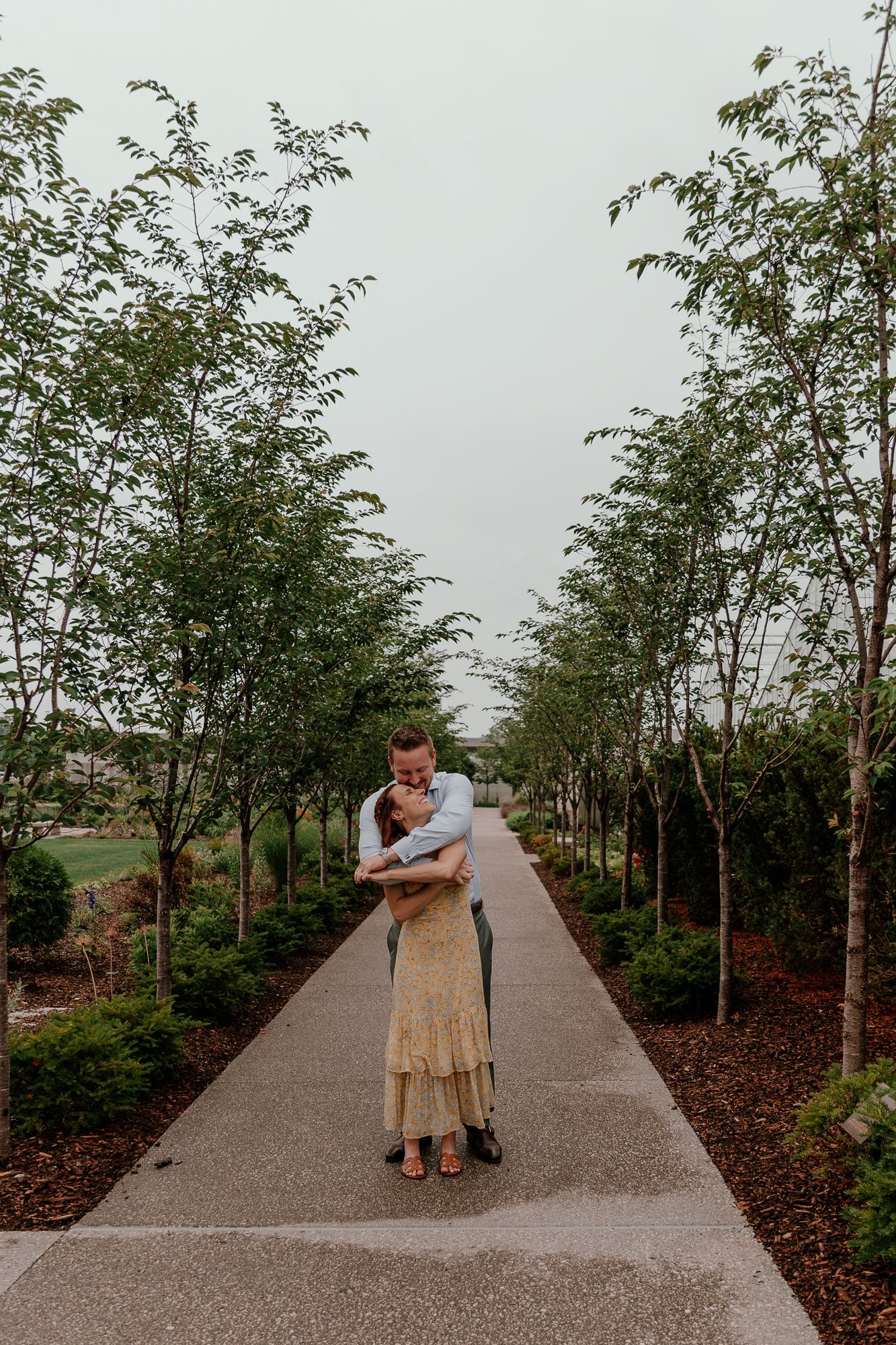 A couple embraces on a tree-lined pathway in a serene garden setting.