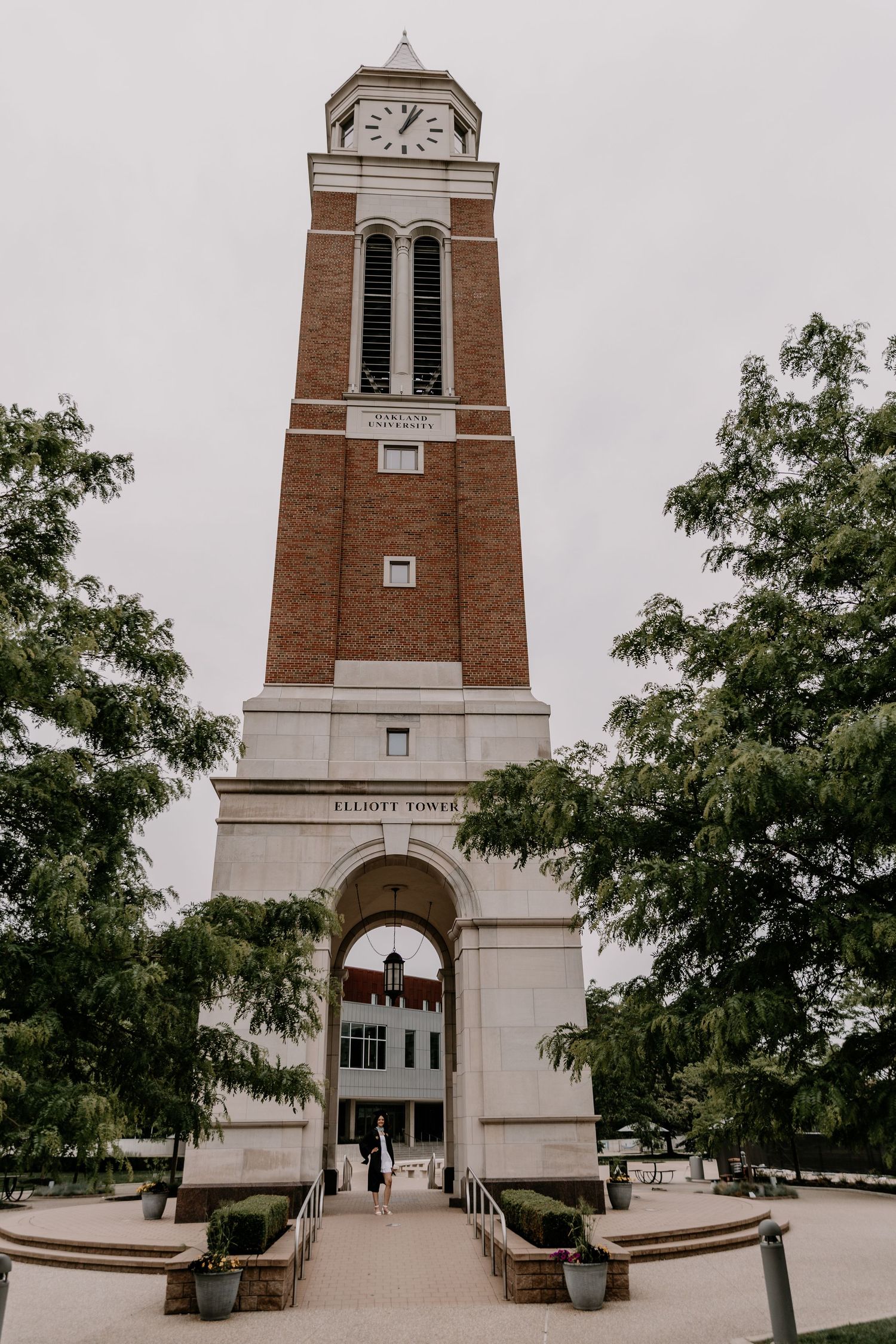 A tall brick clock tower with white architectural details stands at the entrance of a university building framed by trees.