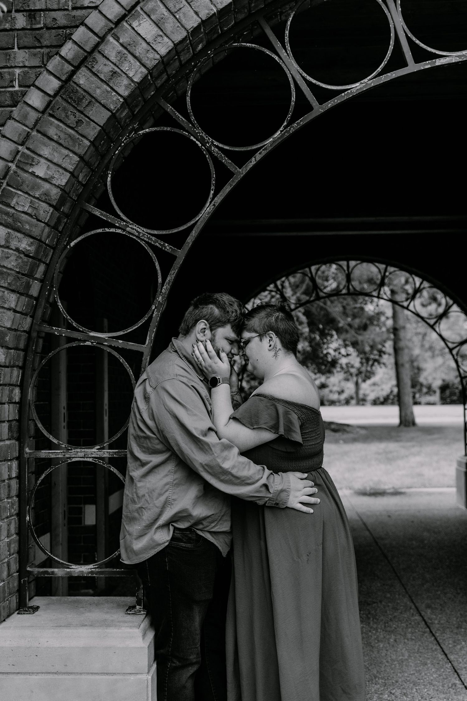 Black and white photo of a couple sharing a romantic moment under a decorative brick archway in a park.